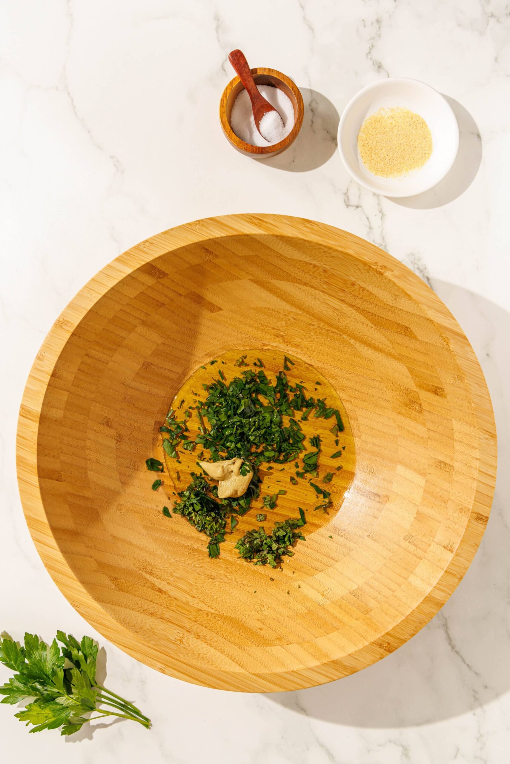 Large salad bowl with olive oil, herbs, and dijon mustard in it. Behind the bowl is two smaller bowls of salt and garlic powder.