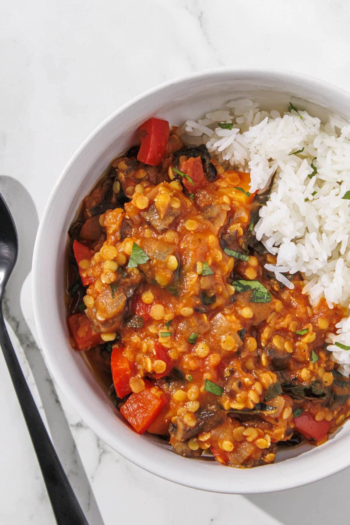 One white bowl filled half with white rice and the other half with the other half with a red lentil, tomato, sweet potato, and spinach mixture. Beside the bowl is a black metal spoon.