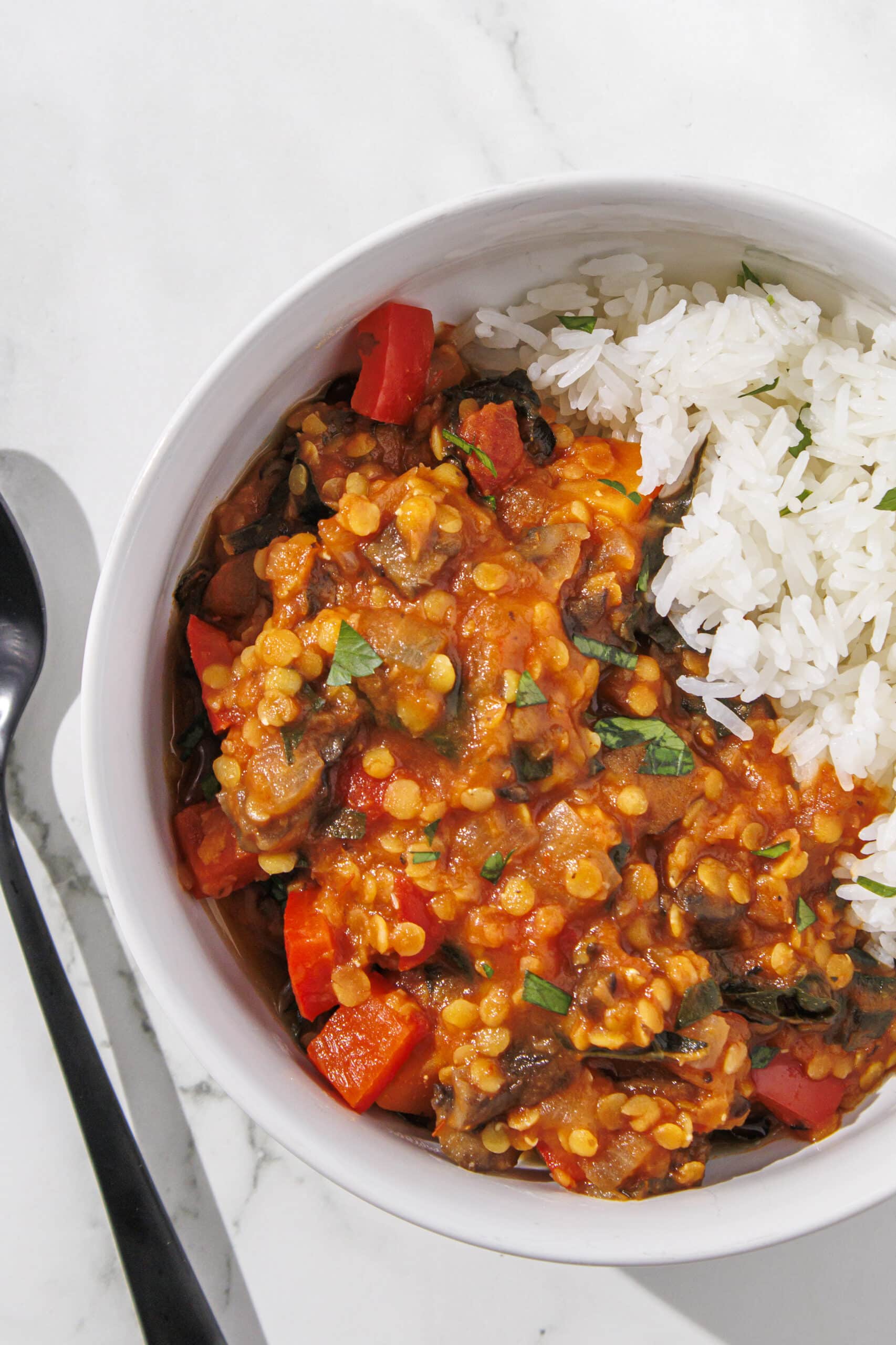 One white bowl filled half with white rice and the other half with the other half with a red lentil, tomato, sweet potato, and spinach mixture. Beside the bowl is a black metal spoon.