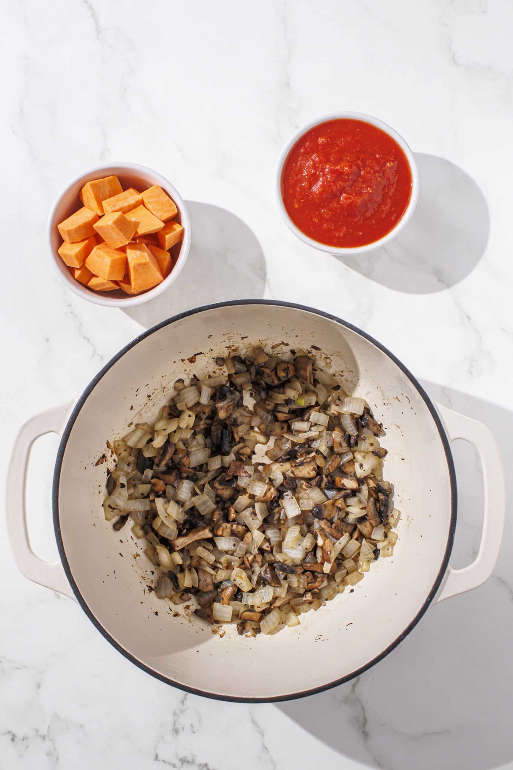 A large pot filled with onion and mushroom sauteed. Beside the pot is two small bowls of raw sweet potato cubes and crushed tomatoes.