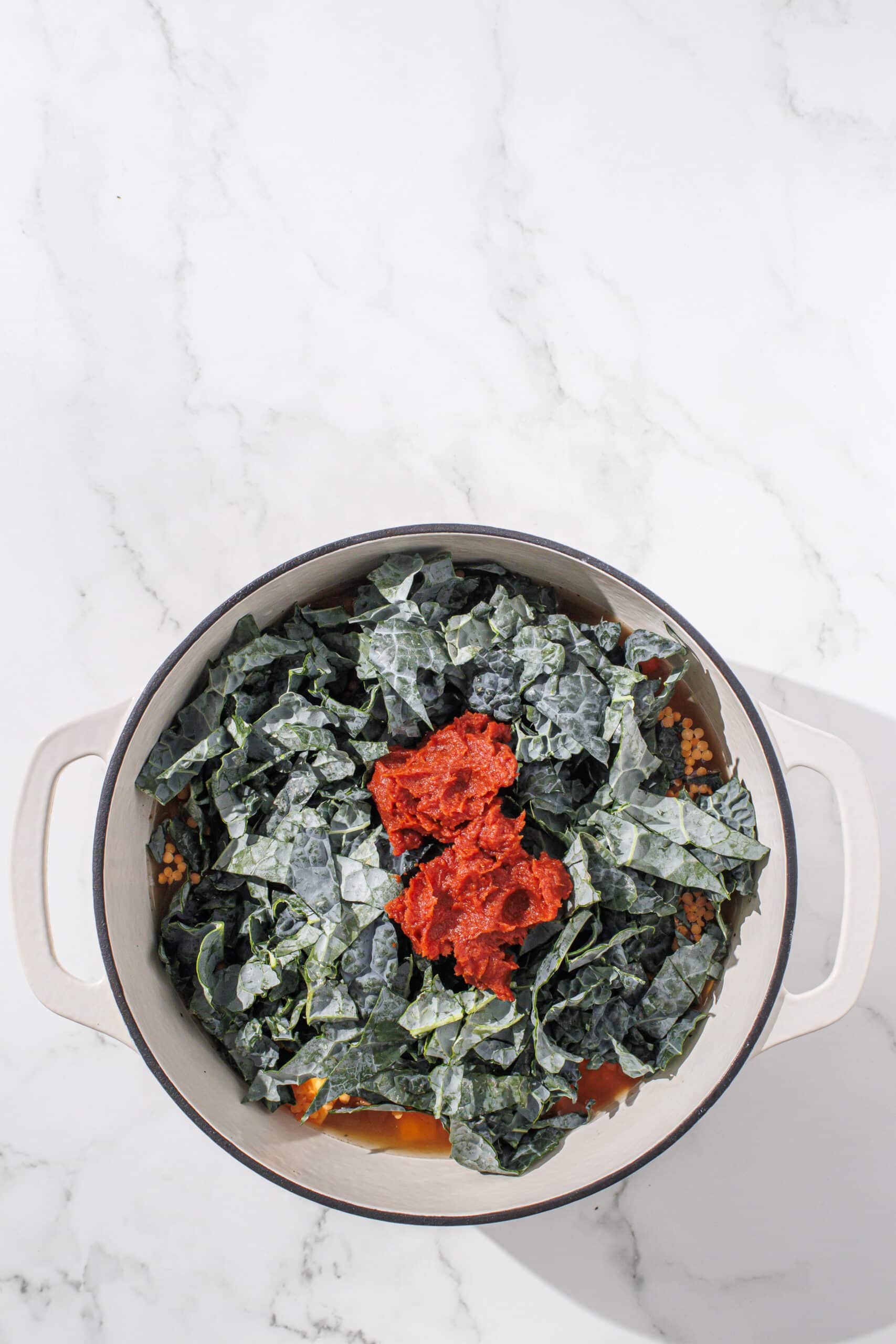 A large white pot filled with nourish pot ingredients under neath topped with raw kale and tomato paste on top ready to be stirred and cooked.