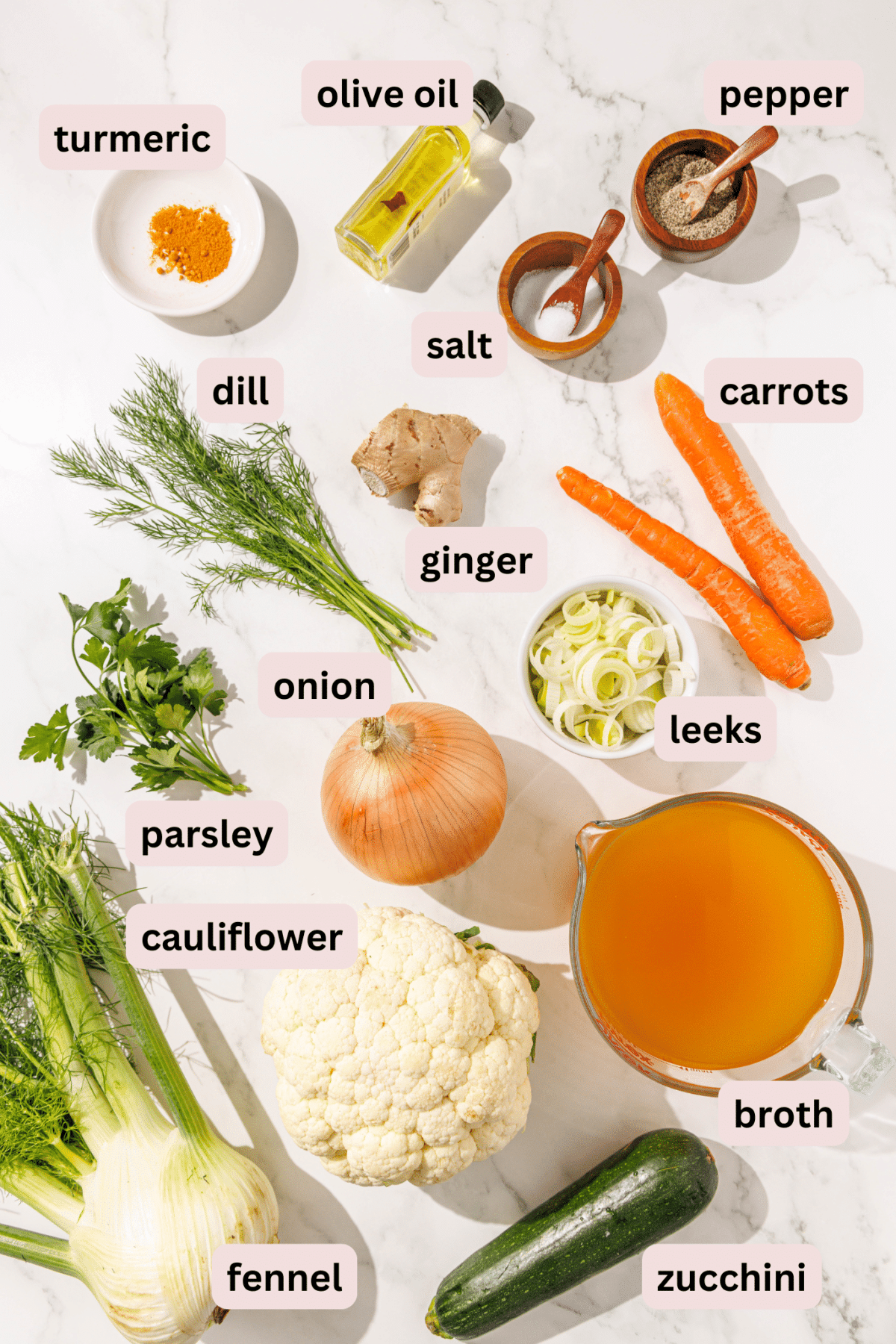 Ingredients needed to make debloat detox vegetable soup laid out on a marble counter and labeled including: turmeric, olive oil, salt and pepper, carrots, ginger, dill, leeks, onion, parsley, cauliflower, broth, fennel, and zucchini.