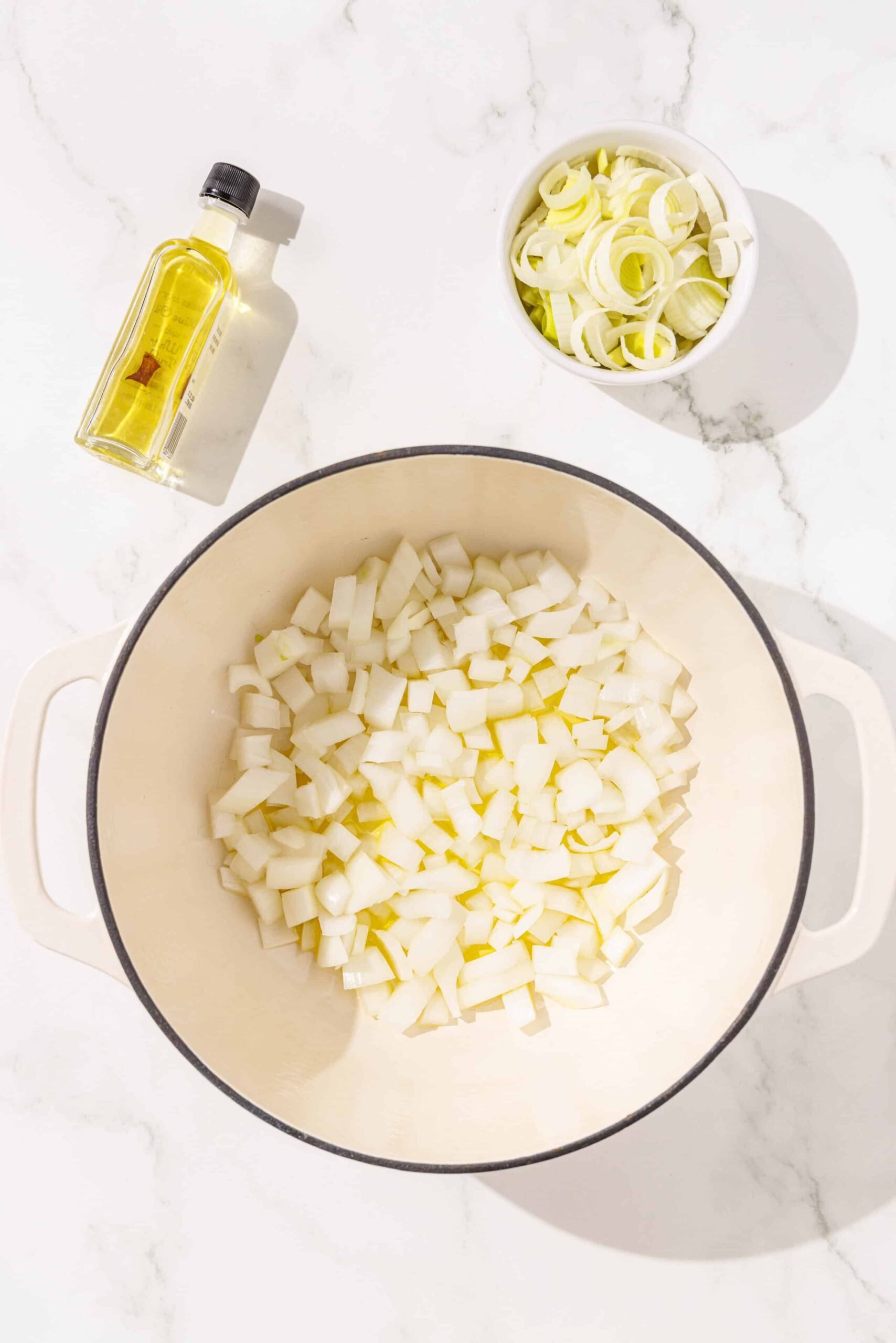 A large pot filled with chopped onions in some olive oil. Beside it is a smaller white bowl of more chopped onions and a small bottle of oil.