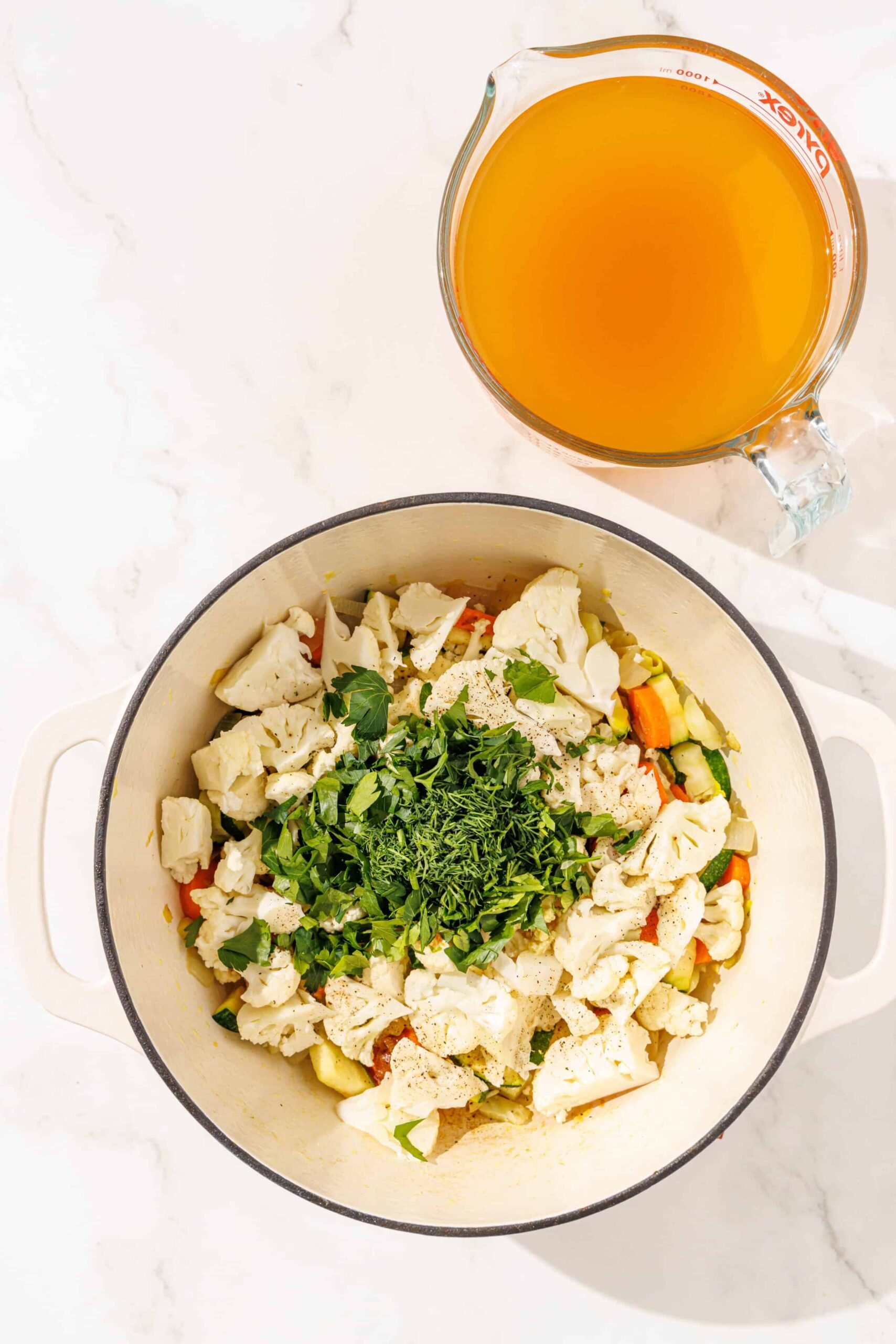 A large soup pot filled with sauteed onions, zucchini, cauliflower, carrots, leeks, and fresh herbs. Beside it is a large glass filled with vegetable broth.