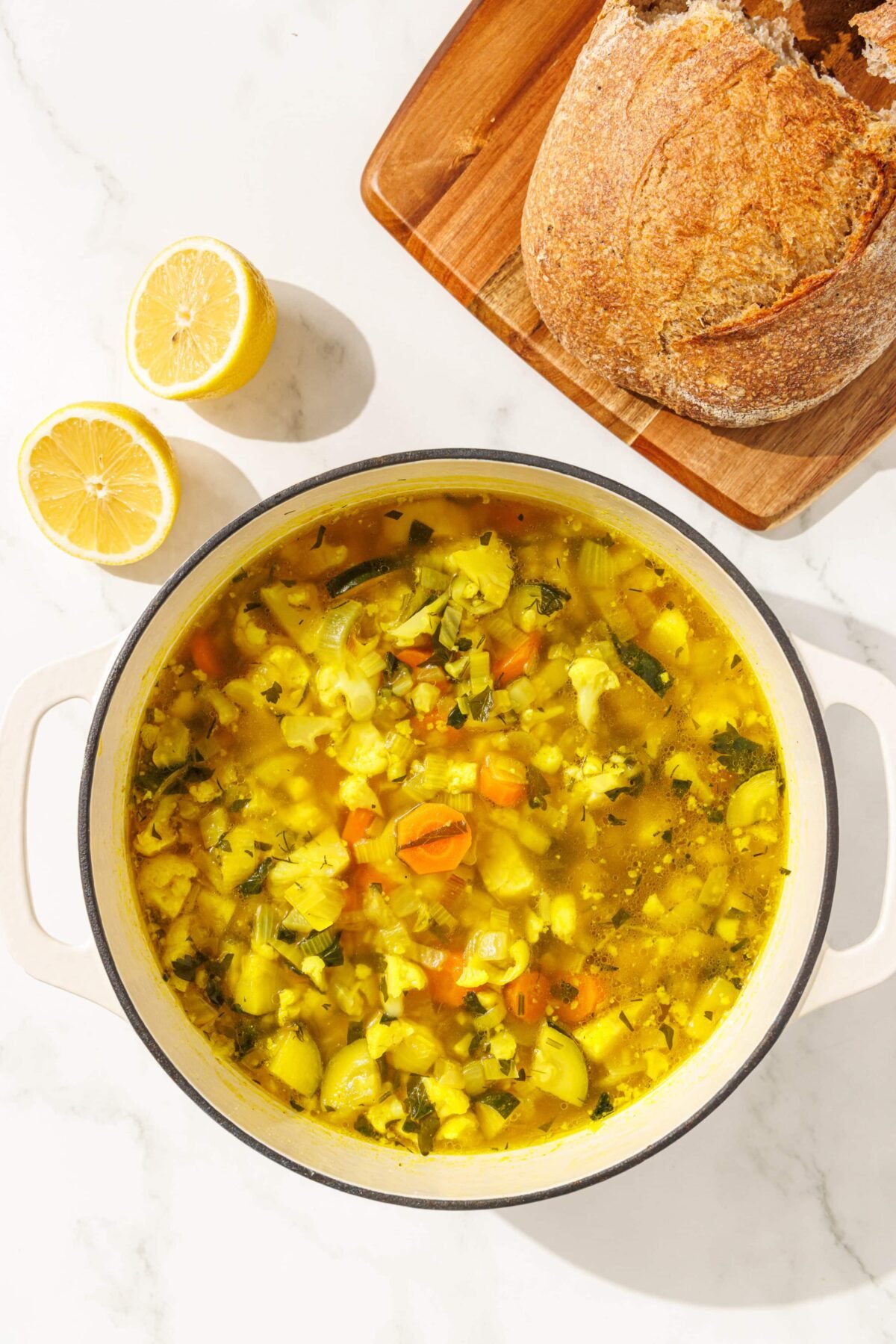 A large soup pot filled with debloat detox vegetable soup. Beside it is a lemon cut in half and a loaf of sourdough bread on a wooden cutting board.