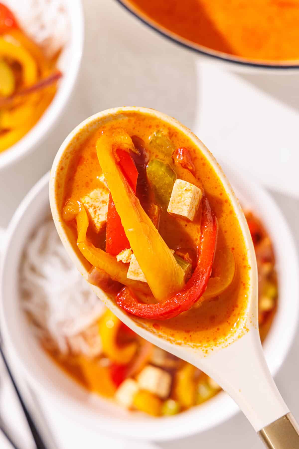 A soup spoon holding a bite of red curry noodle soup with sliced bell pepper and cubes of tofu on it.