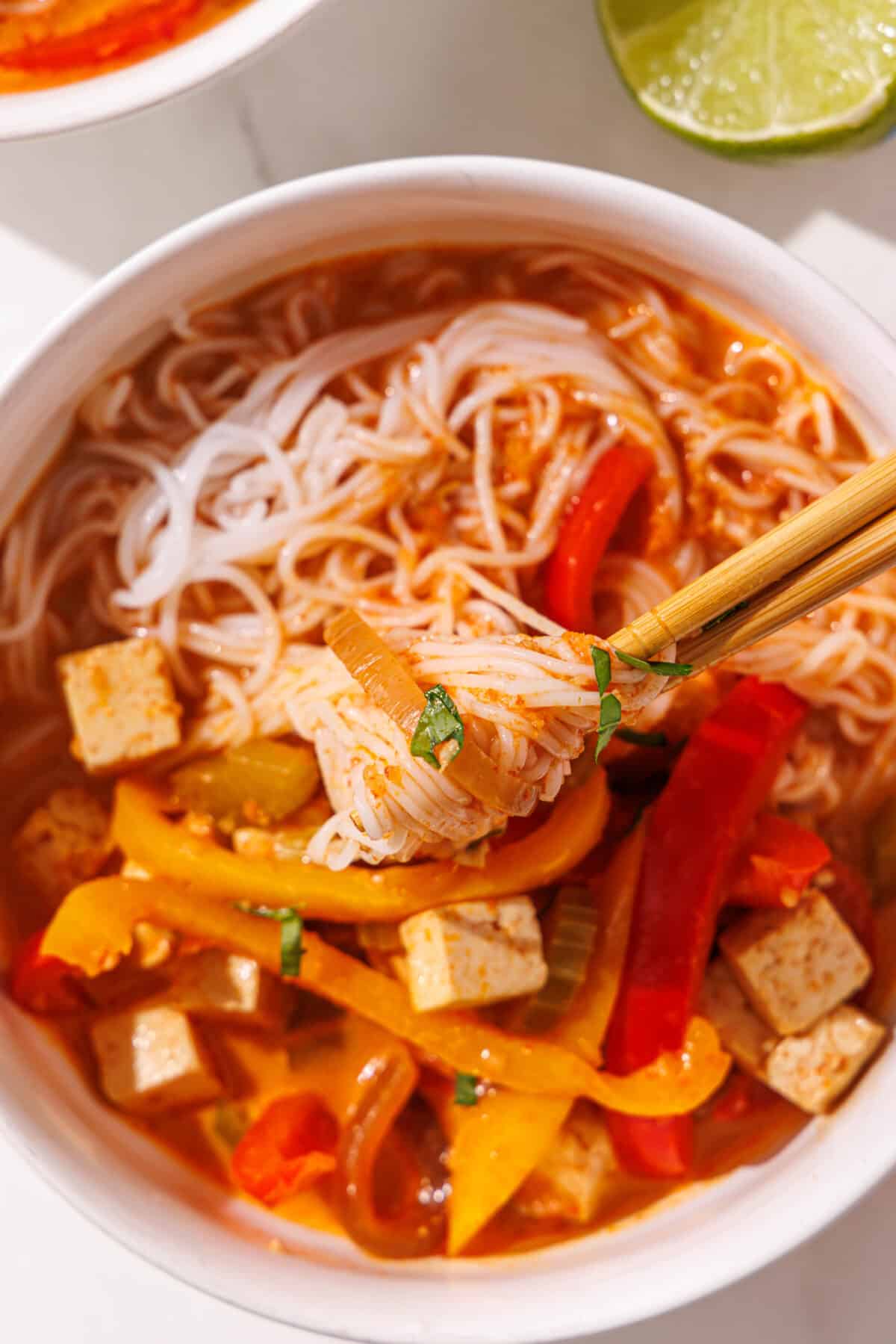A bowl of red curry noodle soup with tofu and bell peppers in it and a set of chopsticks. Beside the bowl is half of a lime.