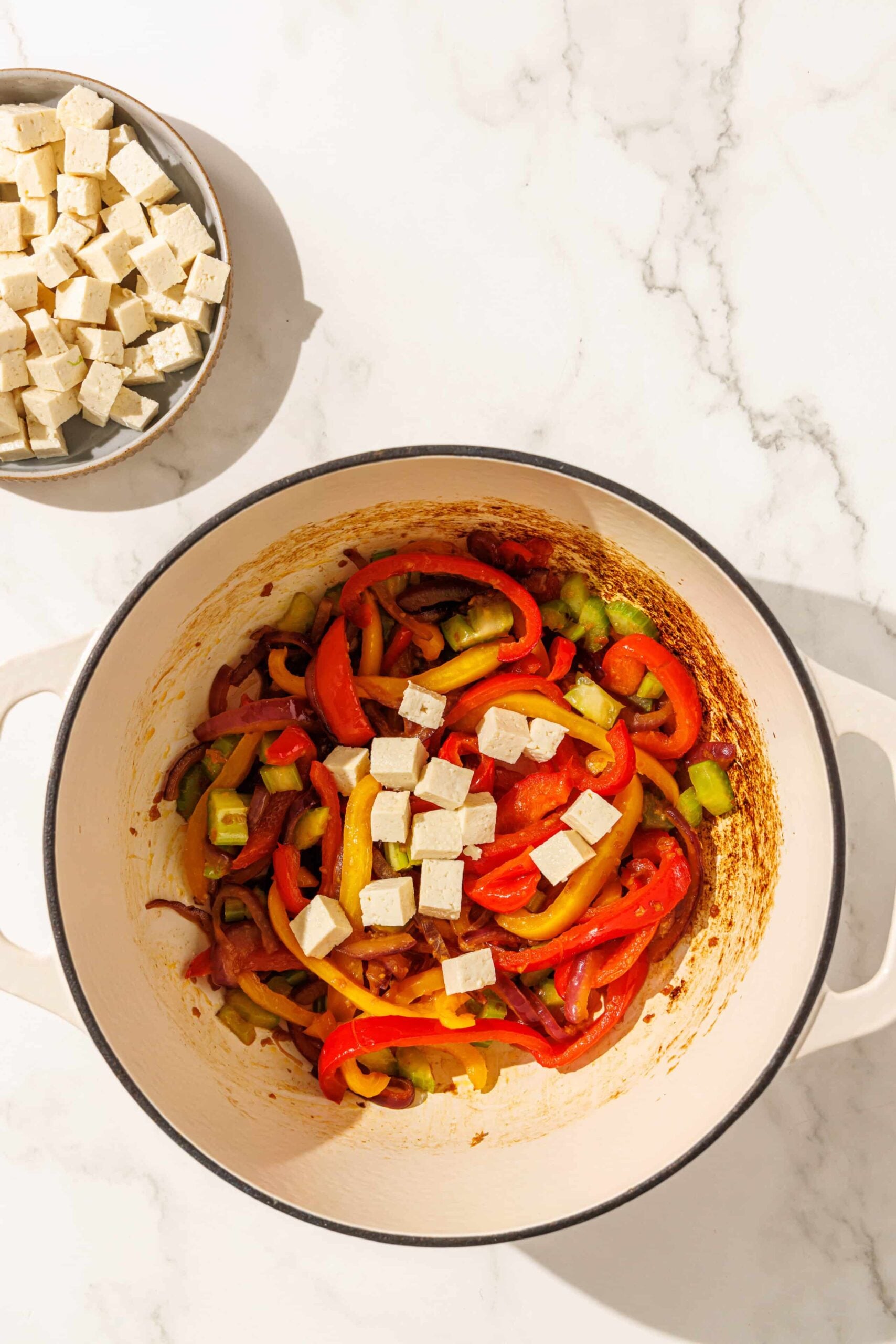 A dutch oven pot filled with sliced peppers and onions and cubes of tofu. Beside the pot is a bowl of tofu cubes.