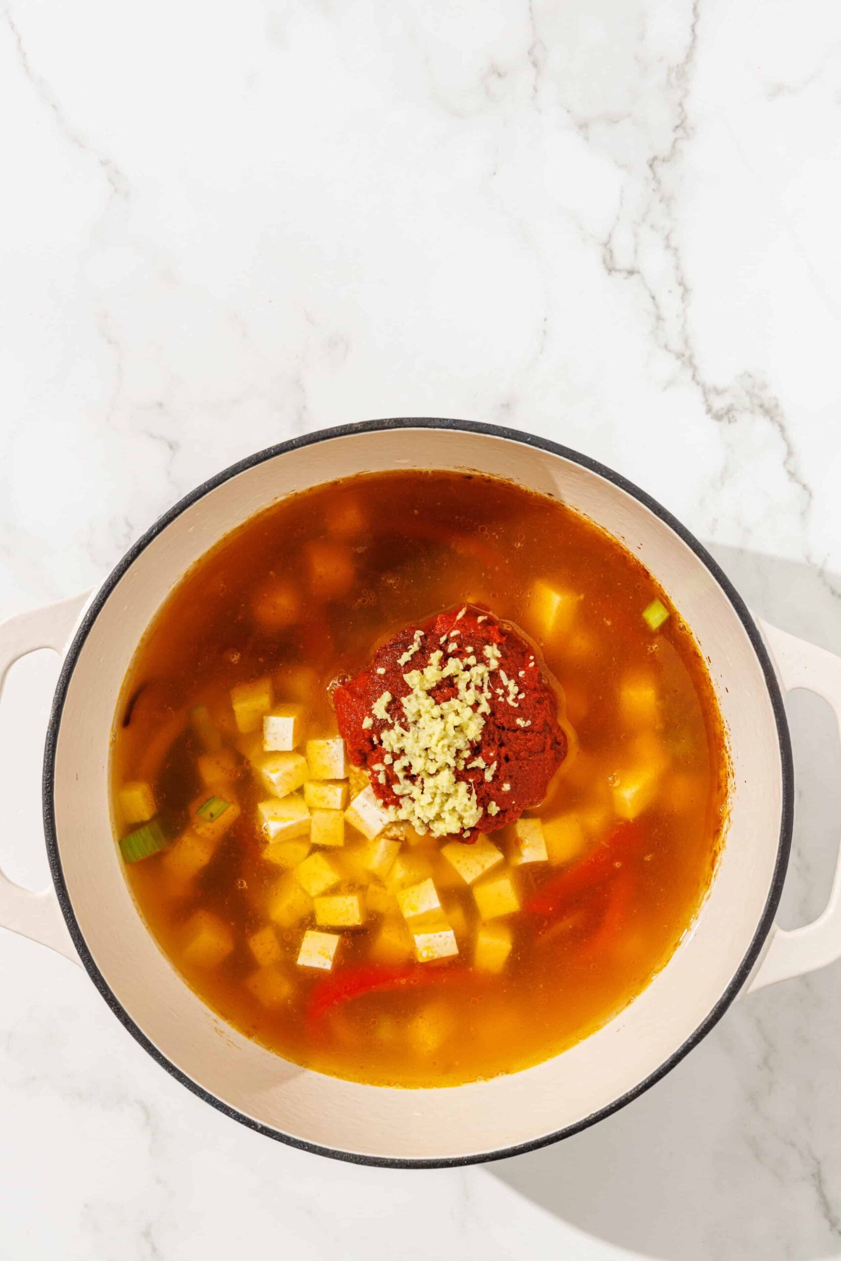 A pot filled with sliced bell peppers, onions, cubes of tofu, red curry paste, ginger, and vegetable broth.
