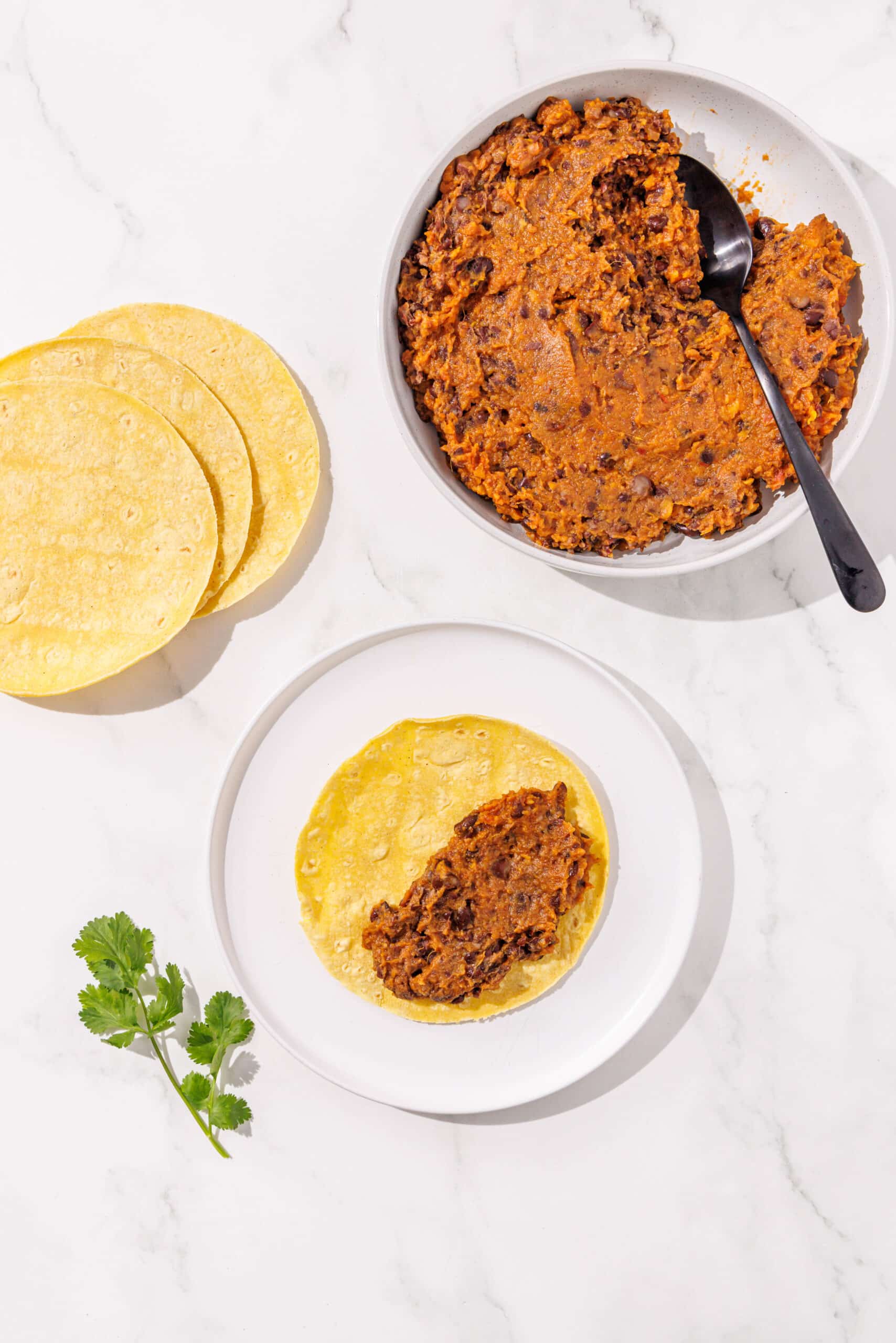 A white bowl filled with bean and sweet potato taco filling and a large black spoon. To the left is a stack of corn tortillas and a white plate with a corn tortilla on it filled with taco filling on the right side. There is a sprig of cilantro beside the white plate.