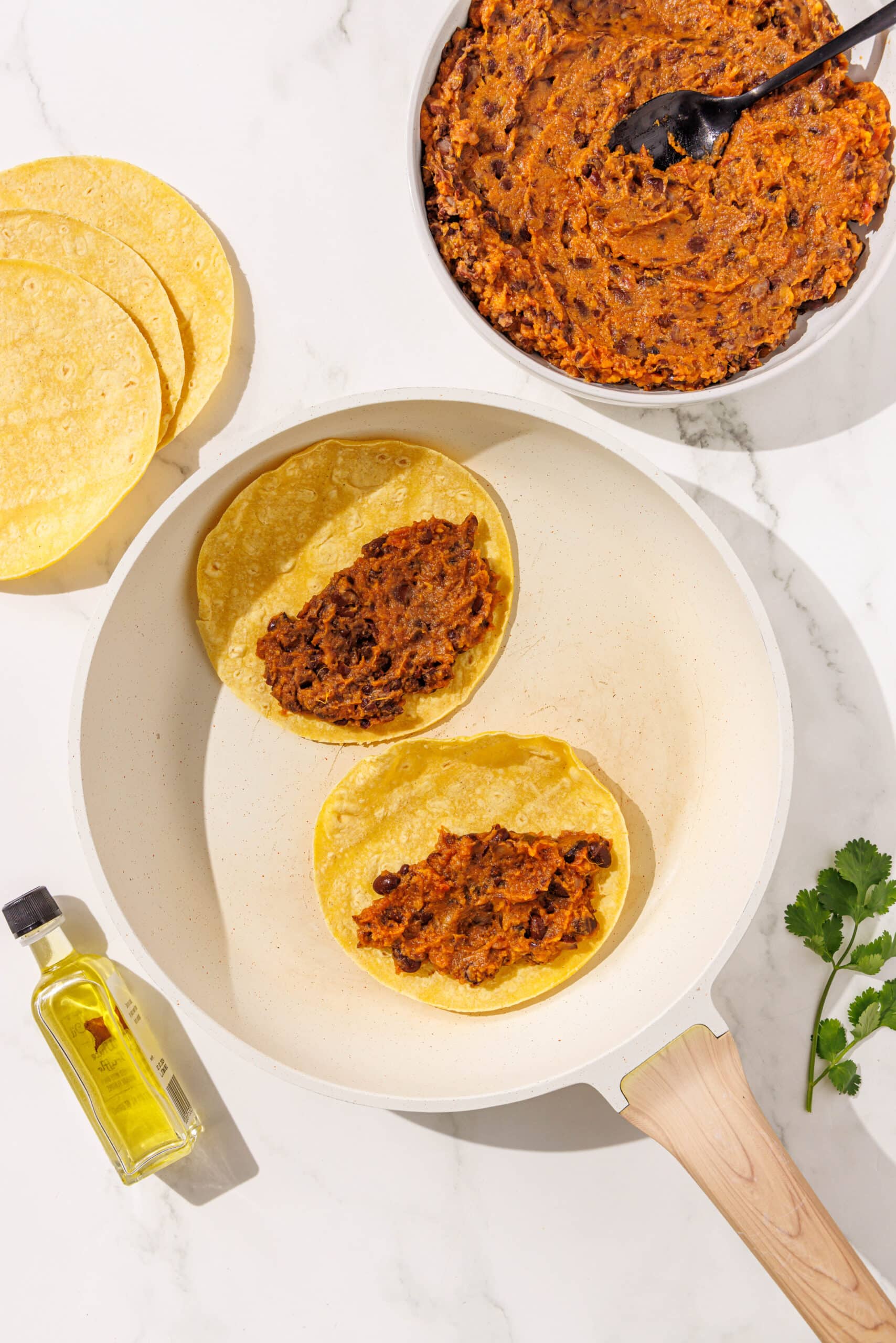 Two bean and sweet potato tacos frying in a small white pan. Around the pan is a small bottle of oil, a stack of corn tortillas, a piece of cilantro, and a large bowl of taco filling with a black spoon in it.