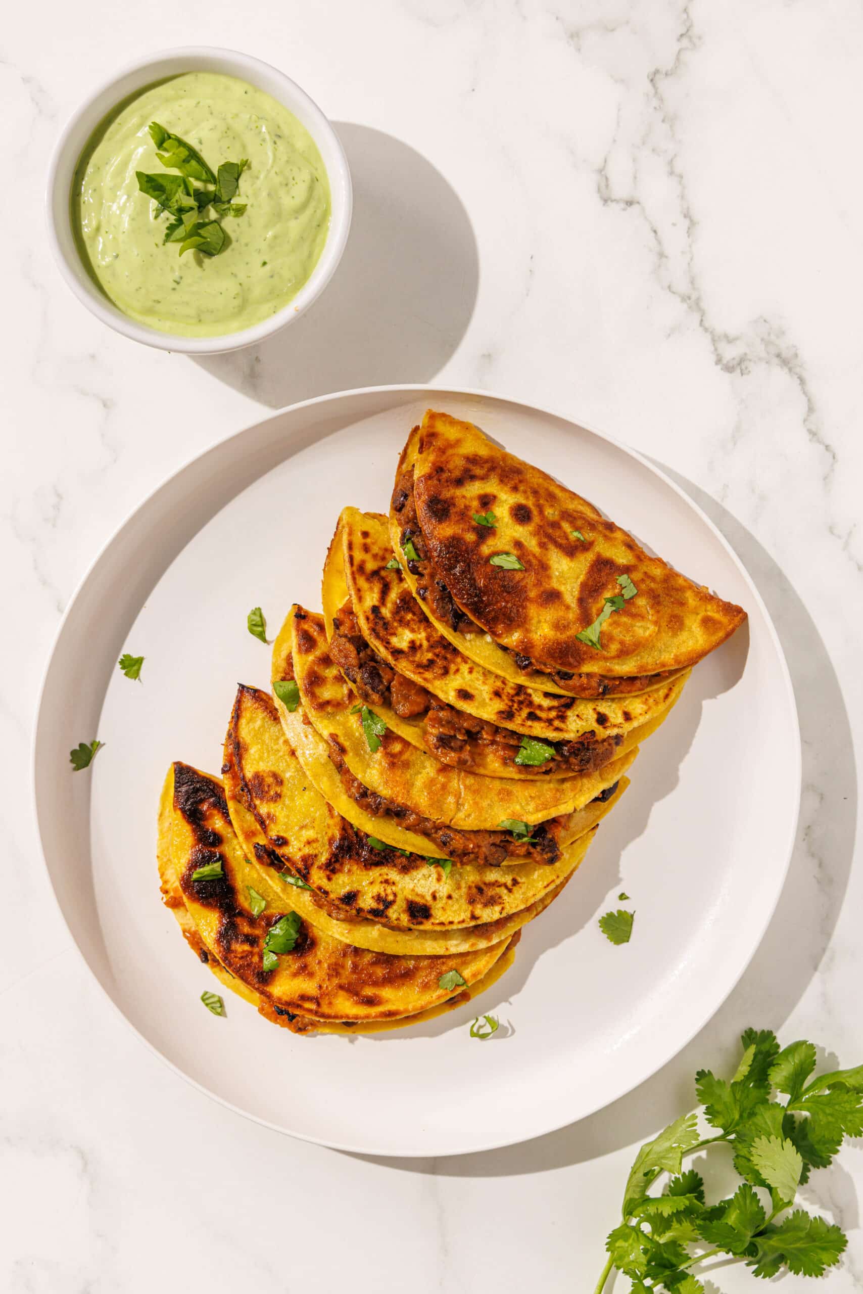 Bean and sweet potato tacos stacked side by side on a white plate garnished with cilantro. To the left is a small white bowl of avocado crema.