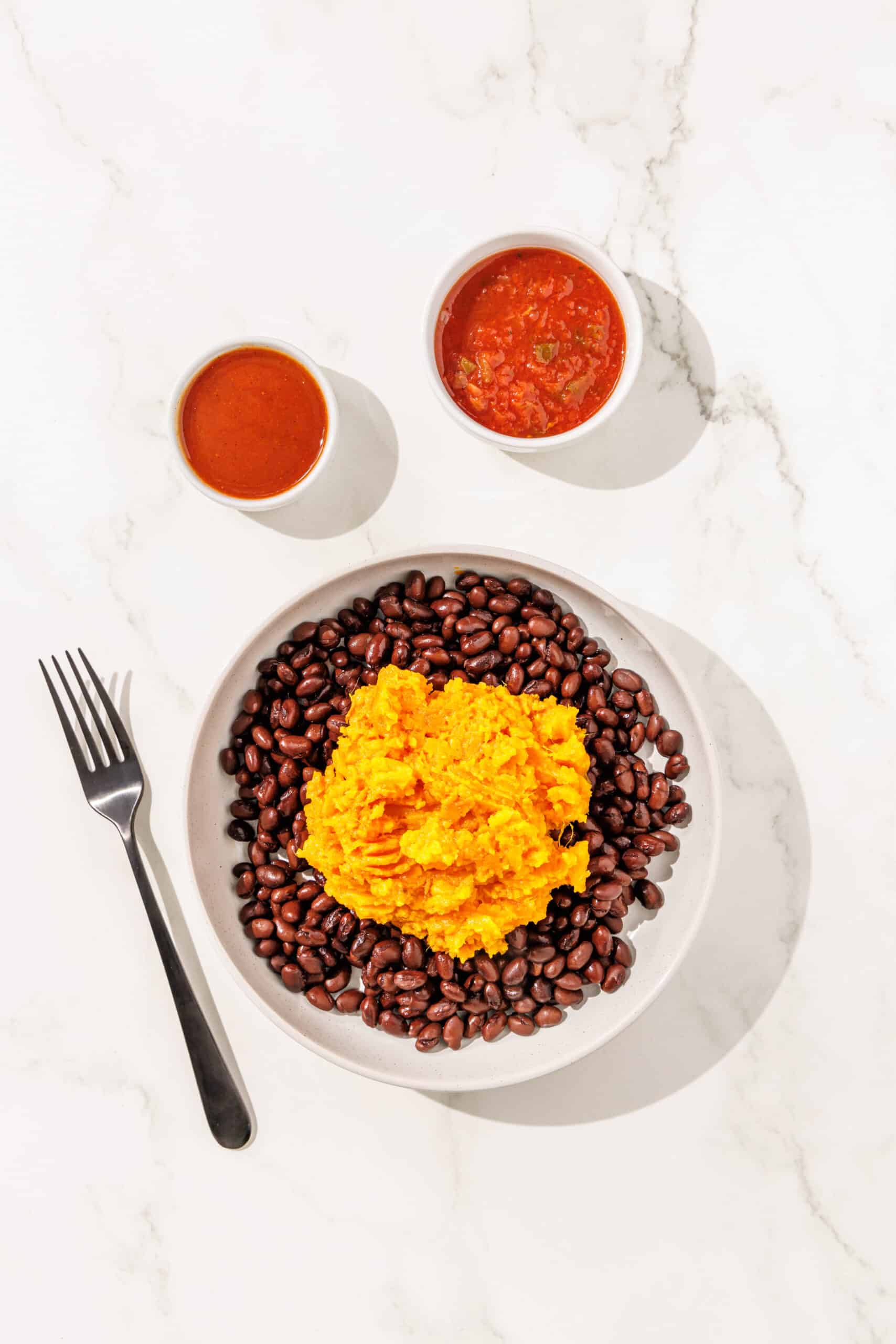 A white bowl of black beans and mashed sweet potato. Beside the bowl is a black fork, and two white bowls of taco sauce and salsa.