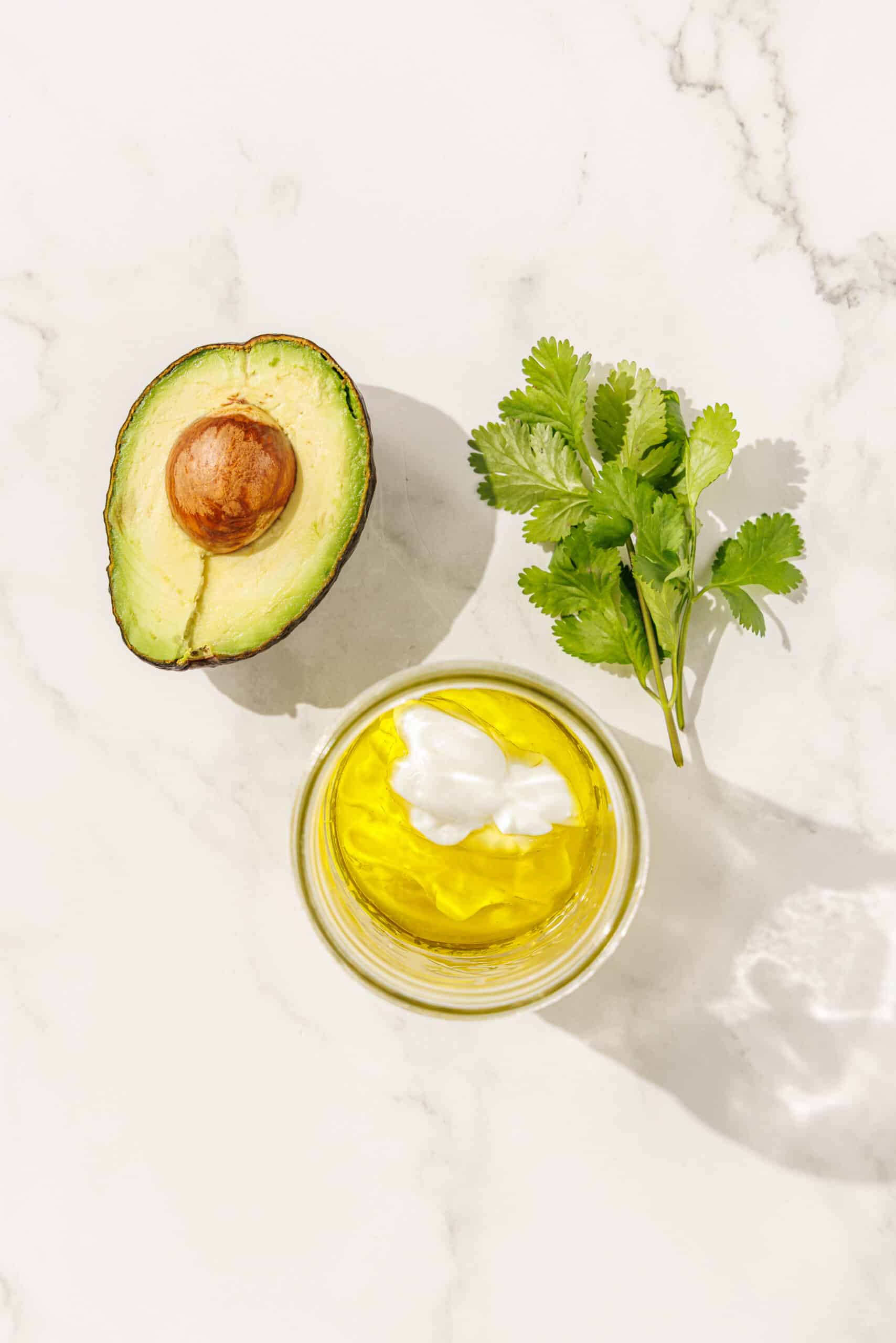A glass jar filled with oil and vegan yogurt. Above the jar is a sprig of cilantro and half of an avocado.
