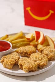 A plate of tofu tenders, baked fries, apple slices, and a small cup of ketchup. In the background is a mcdonalds box.