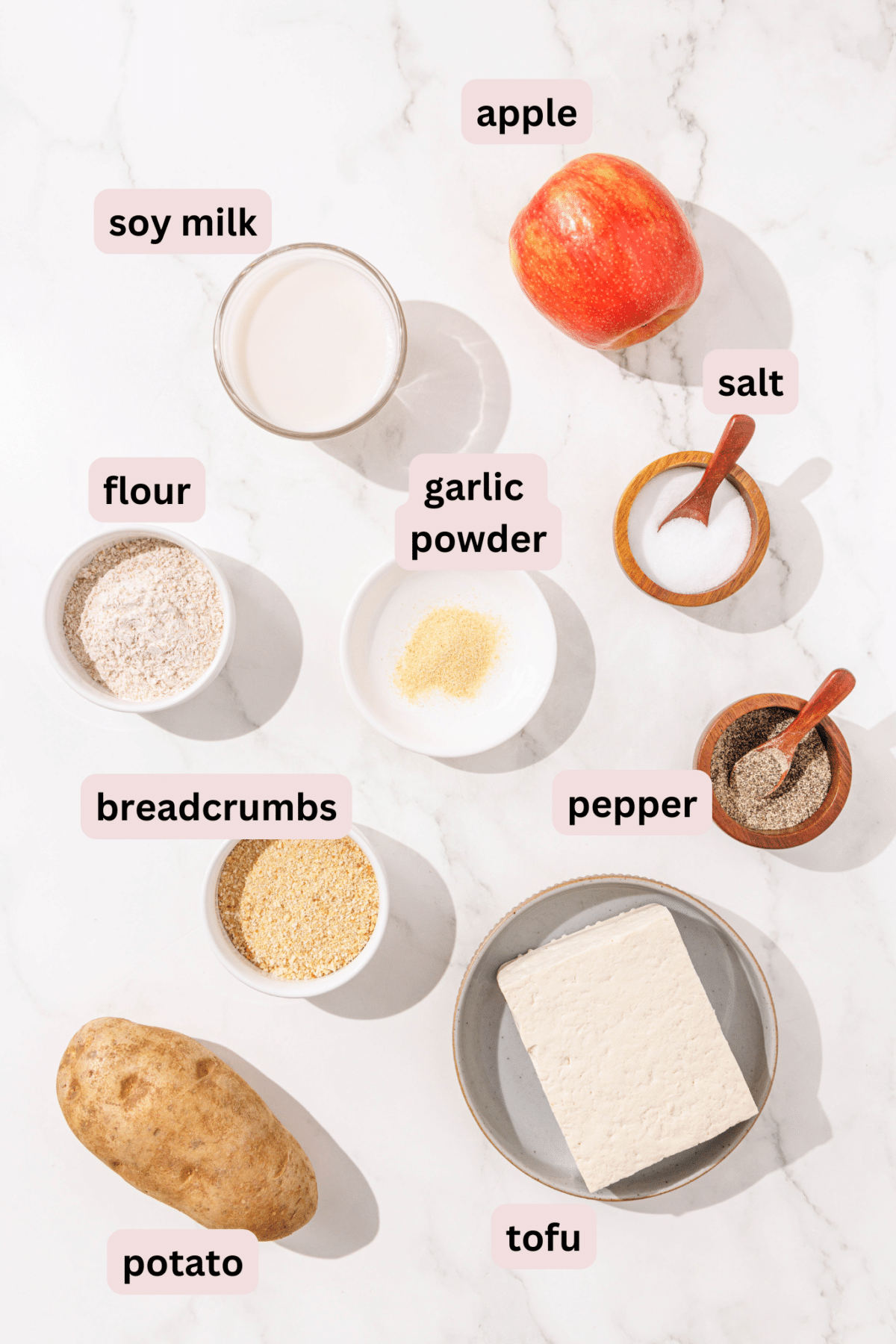 Ingredients laid out on a counter and labeled including: apple, soy milk, salt, garlic powder, flour, pepper, breadcrumbs, tofu, and a potato.