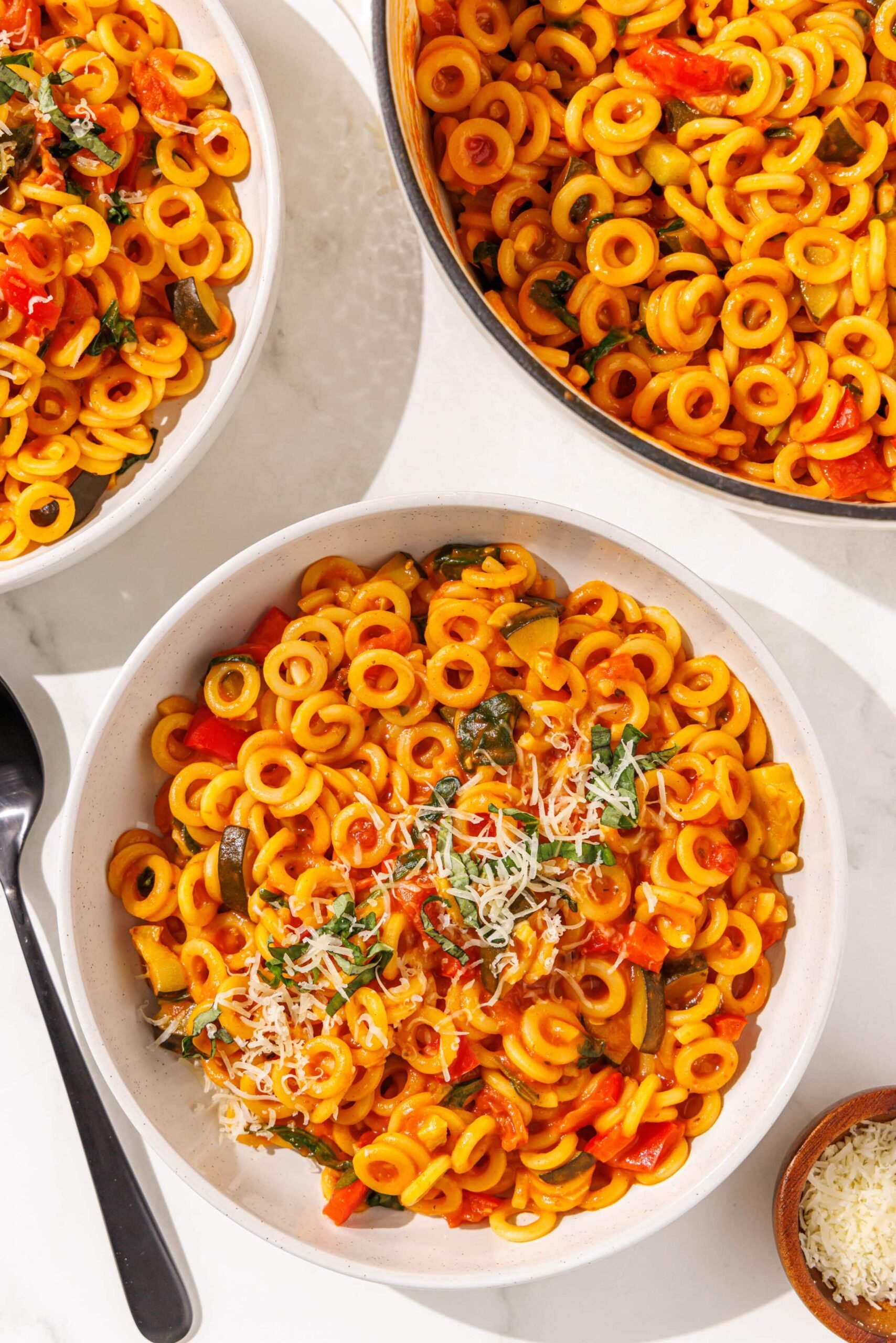 Two white bowls of one pot pasta alongside one large pot of one pot pasta, a small wooden bowl of parmesan, and a black spoon.