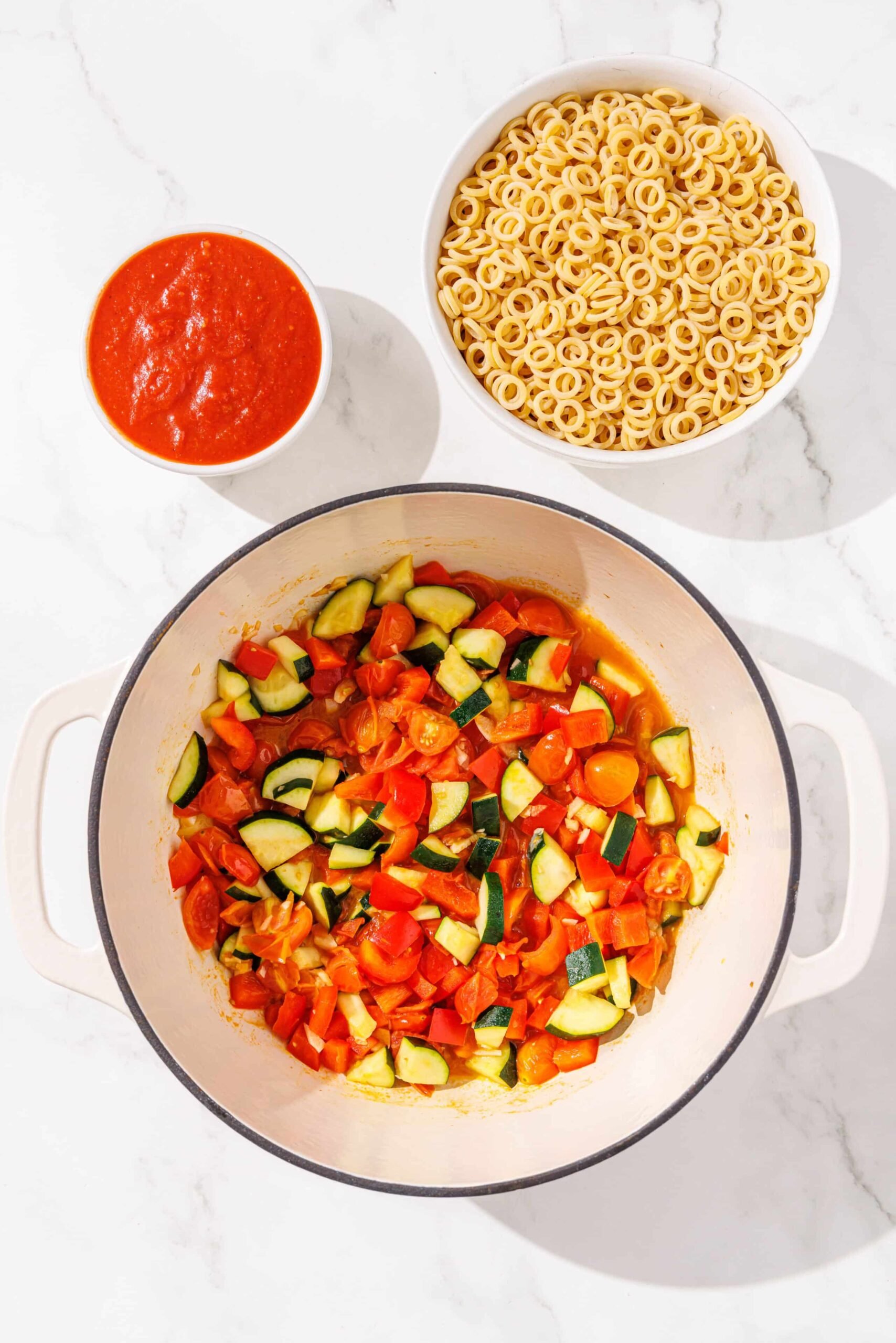 Sauteed zuchinni and bell pepper in a large pot with small white bowls of pasta and marinara beside it.