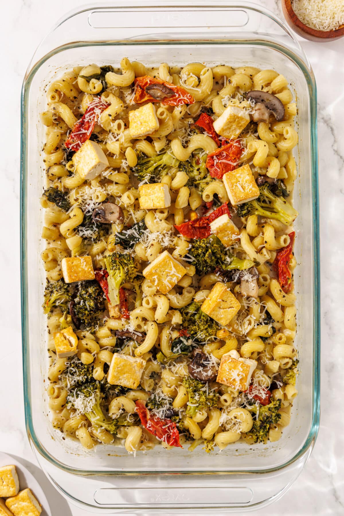 A large glass dish filled with one pan tuscan pasta. A small bowl in the bottom left corner of tofu and another small wooden bowl in the top right corner of vegan parmesan.