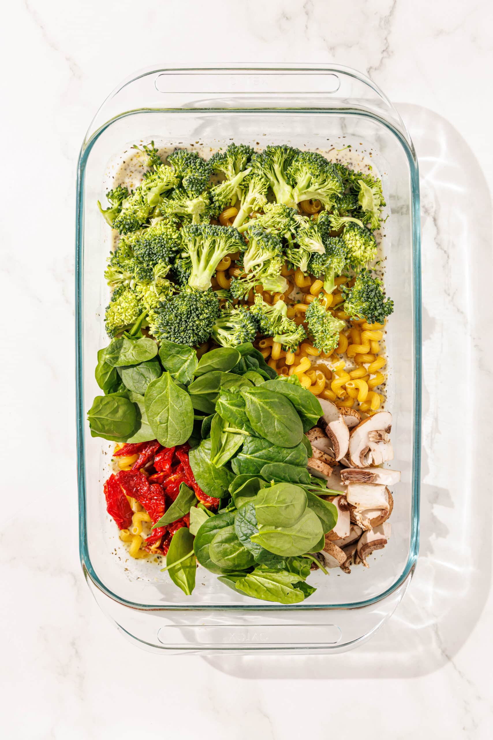A 9x13 glass baking dish filled with tuscan pasta sauce, uncooked noodles, broccoli, spinach, mushrooms, and sundried tomatoes all ready to be baked.