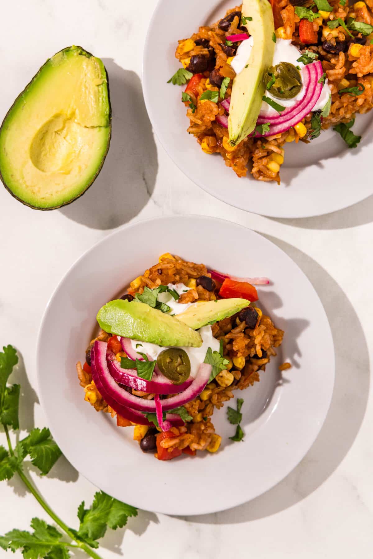 Two white plates each with a serving of one pan taco casserole on them topped with a slice of avocado, pickled onion, jalapeno and cilantro. Beside the plates is a sprig of cilantro and half of an avocado.