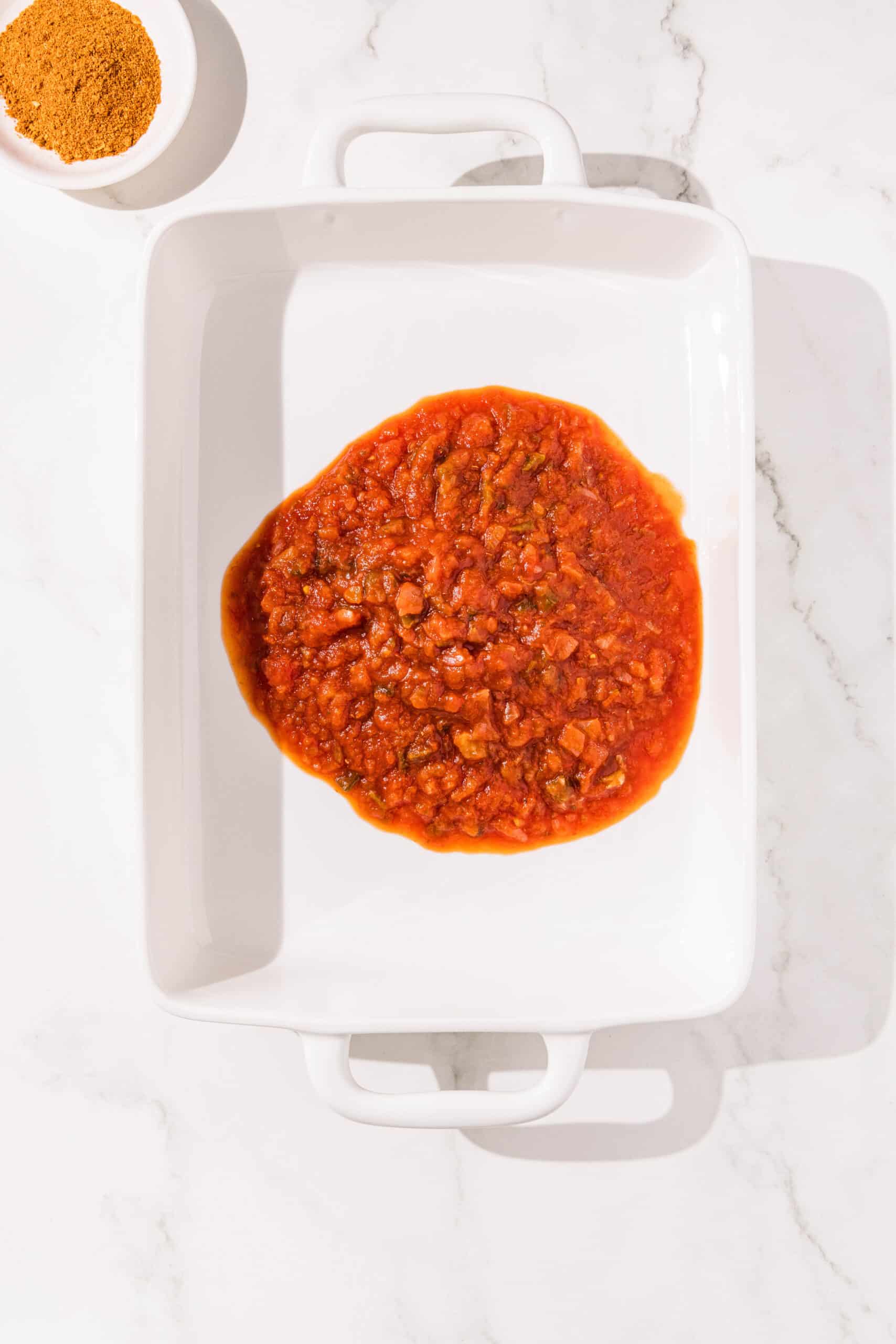 A white casserole dish filled with salsa. Above it is a small white bowl of taco seasoning.