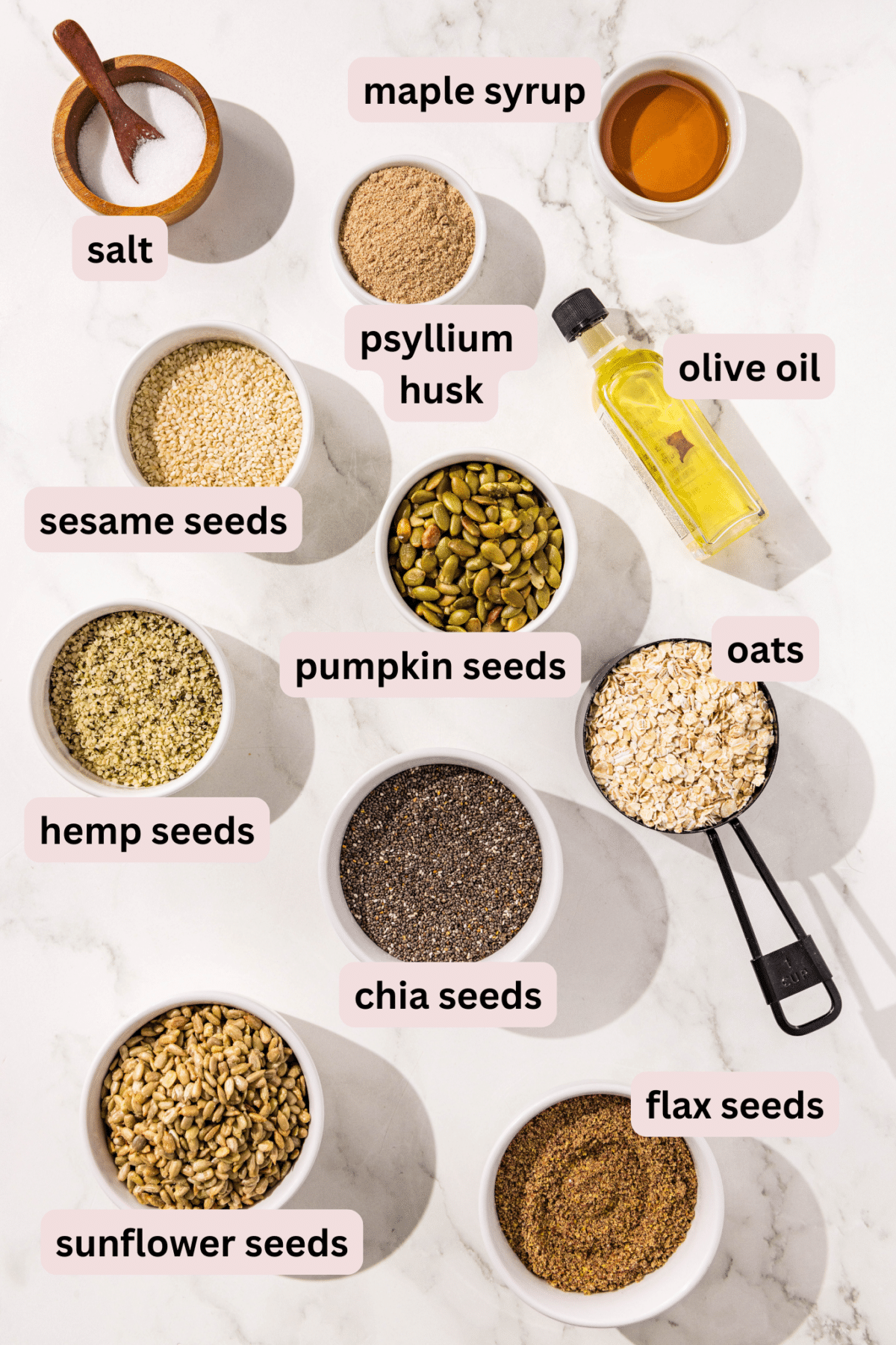 Ingredients needed to make seedy bread laid out on the counter and labeled including: salt, maple syrup, psyllium husk, olive oil, pumpkin seeds, sesame seeds, oats, hemp seeds, chia seeds, flax seeds, and sunflower seeds.
