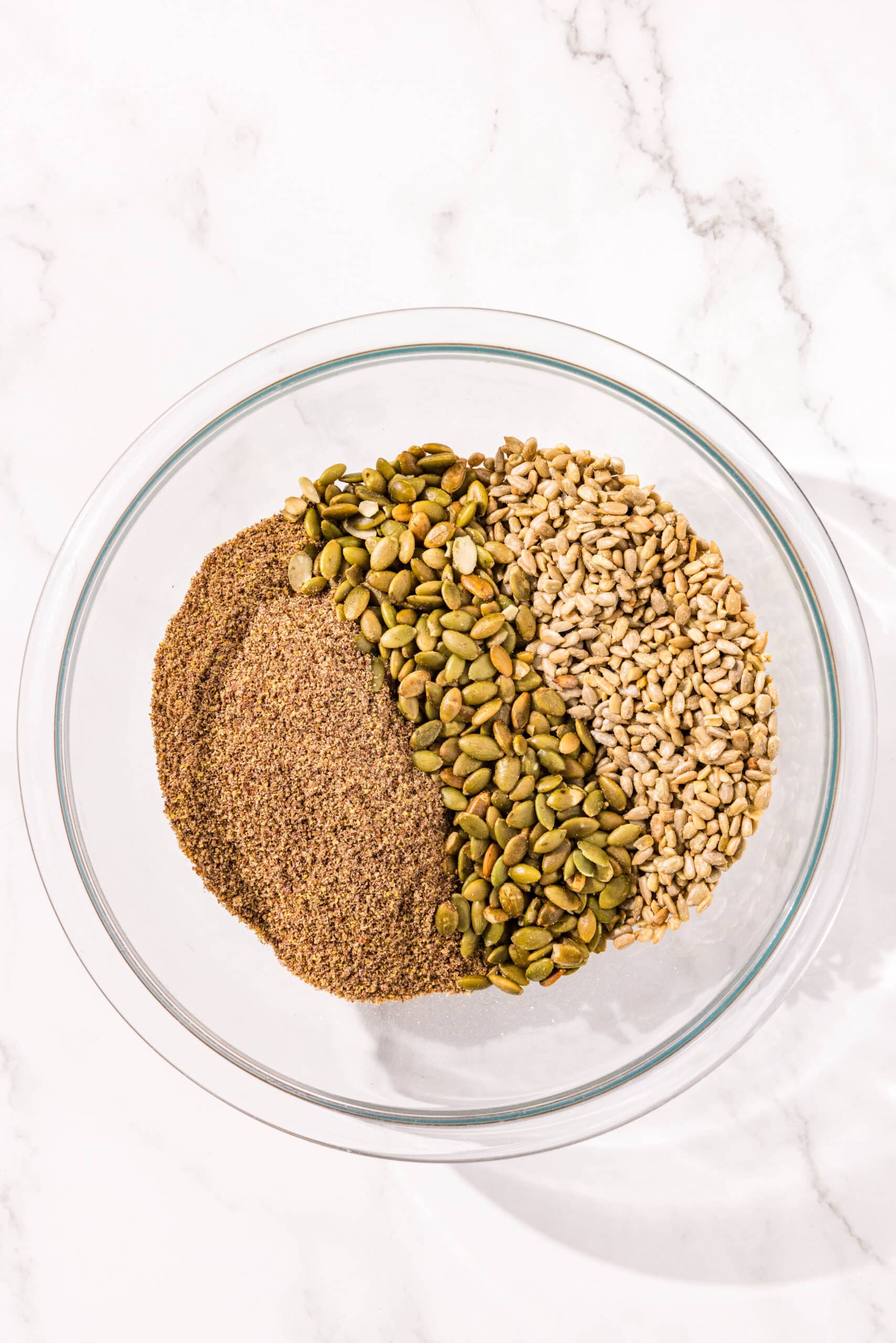 A large glass bowl of pumpkin seeds, sunflower seeds, and flax seeds.