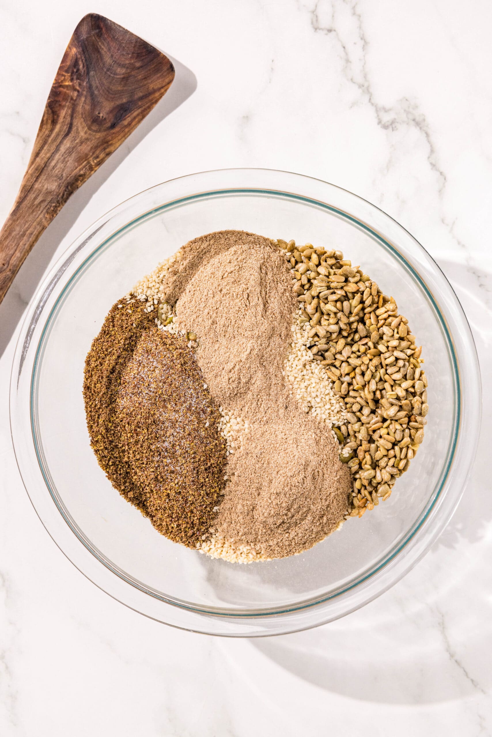 A glass bowl of sunflower seeds, pumpkin seeds, hemp seeds, psyllium husk powder, and flax seeds with a wooden spoon laying beside the bowl.