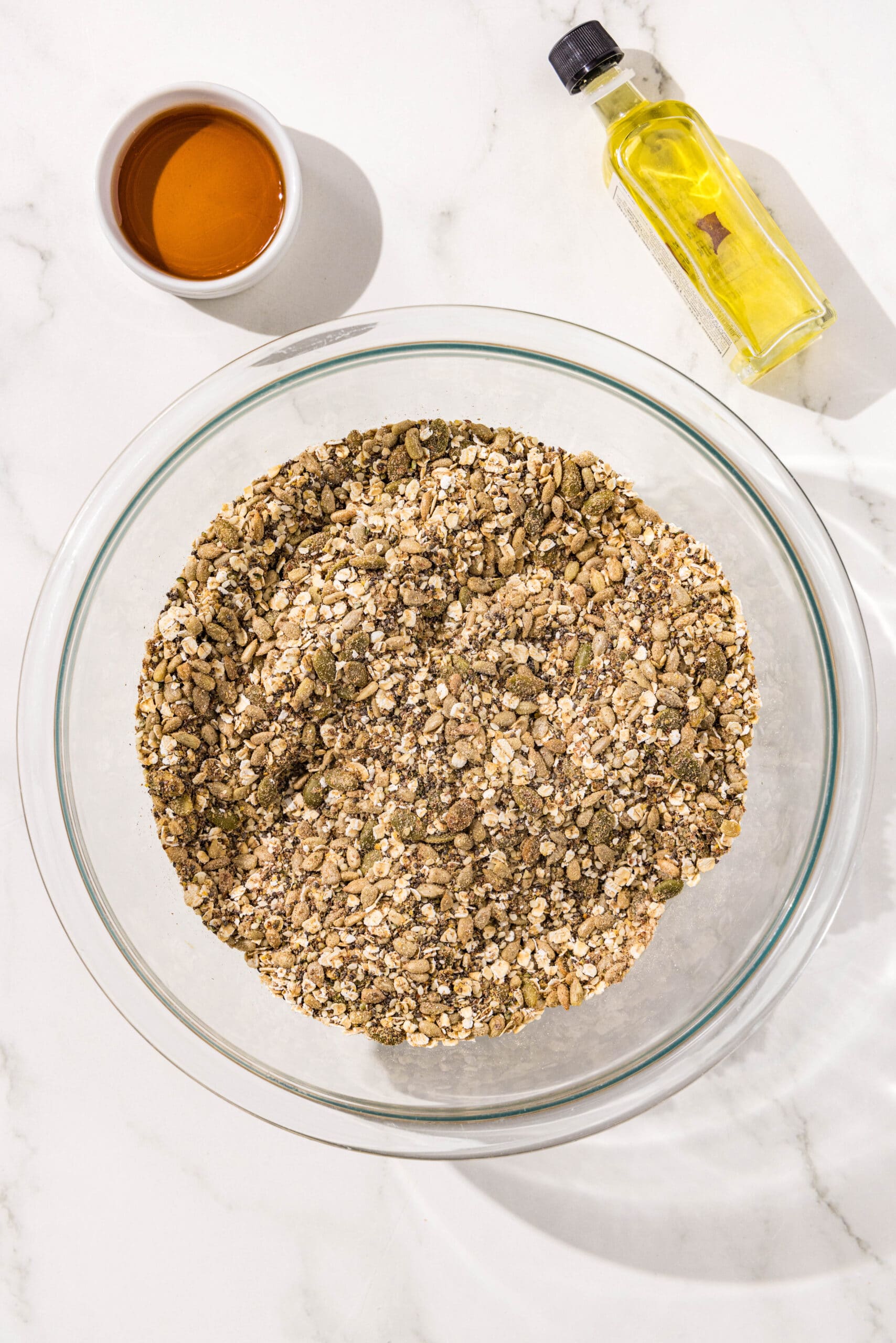 All of the dry ingredients for seedy bread mixed together in a large glass bowl with a small white bowl of maple syrup and small bottle of olive oil beside the bowl.