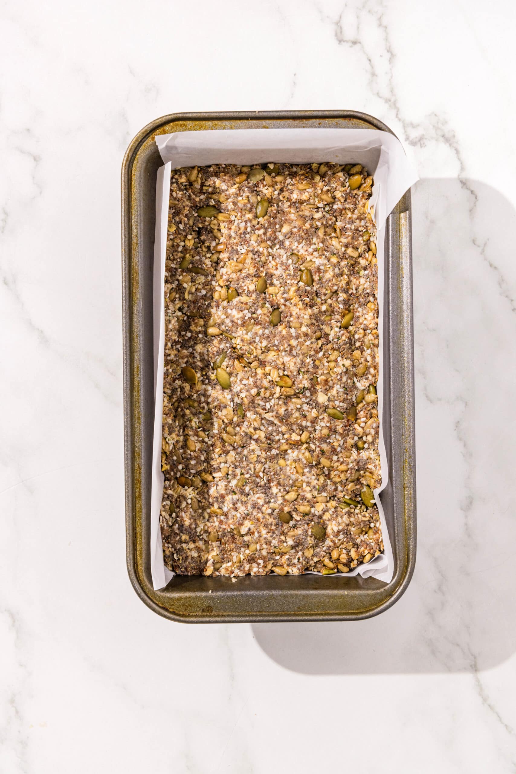 Seedy bread dough in a loaf pan lined with parchment paper ready to be baked.
