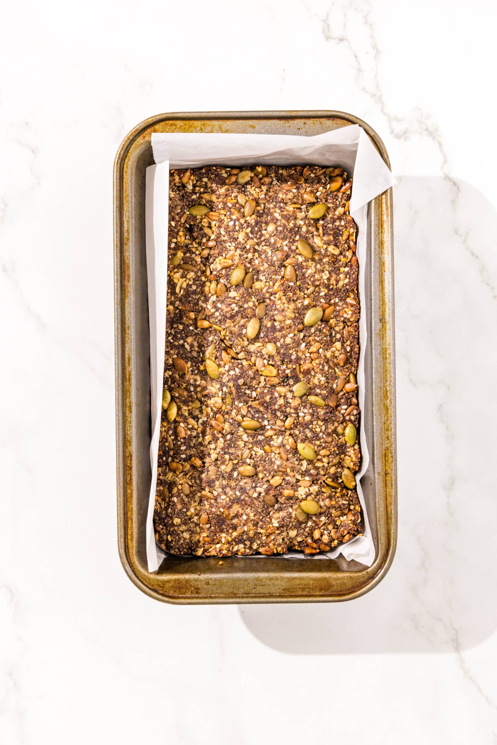 A baked loaf of seedy bread in a loaf pan lined with parchment paper.