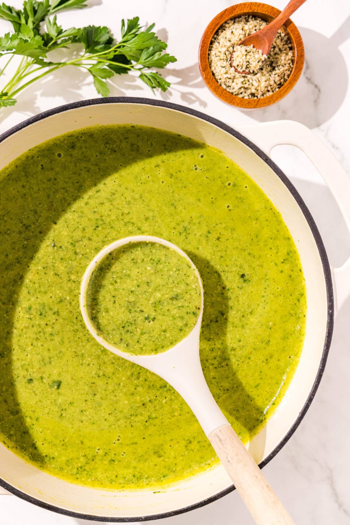 A pot filled with the green goddess soup and a soup spoon inside of the pot. On the side there are hemp seeds in a small bowl and some sprigs of parsley that can be used as garnishes.