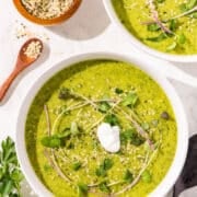 Two bowls of green goddess soup topped with micro greens and sour cream. A small bowl of hemp seeds on the side with a little spoon.