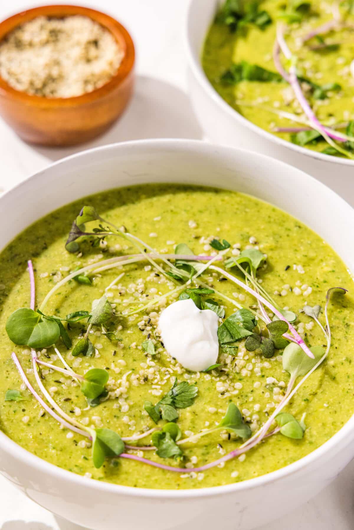 A green soup in a white serving bowl with micro greens, sour cream, and hemp seeds.