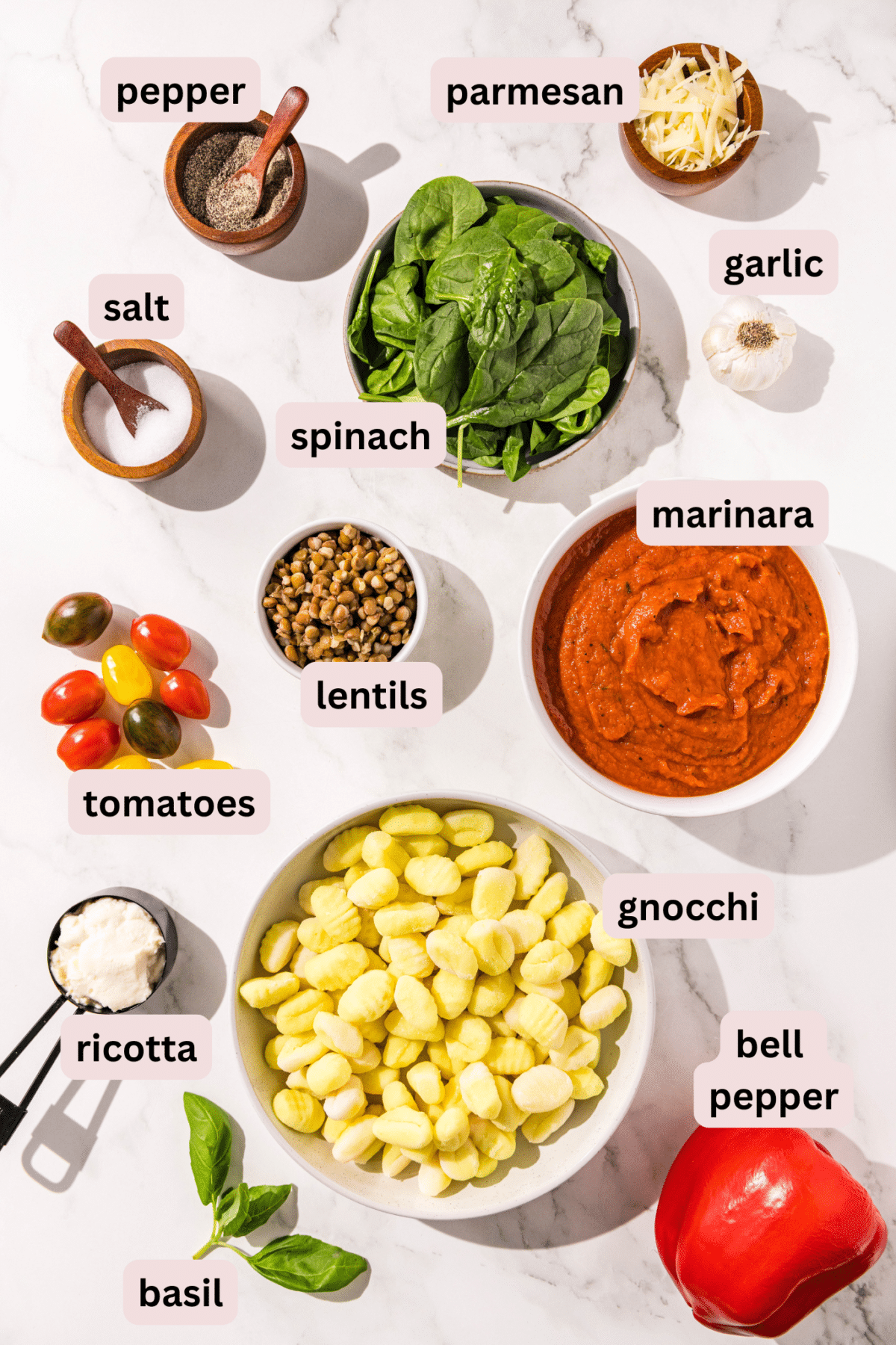 Each ingredient is put out on a table to see what comprises this one pan gnocchi. you can see a bowl of gnocchi, ricotta in a measuring cup, basil, a red bell pepper, marinara in a bowl, cherry tomatoes, lentils in a bowl, salt, pepper, spinach, garlic and parmesan.