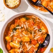 There are two white bowls filled with the One Pan Gnocchi dish garnished with parmesan and basil. Both bowls have a black spoon in them. There is also a small bowl next to the gnocchi bowls filled with parmesan cheese. For aesthetic, there is basil and tomatoes surrounding the bowls.