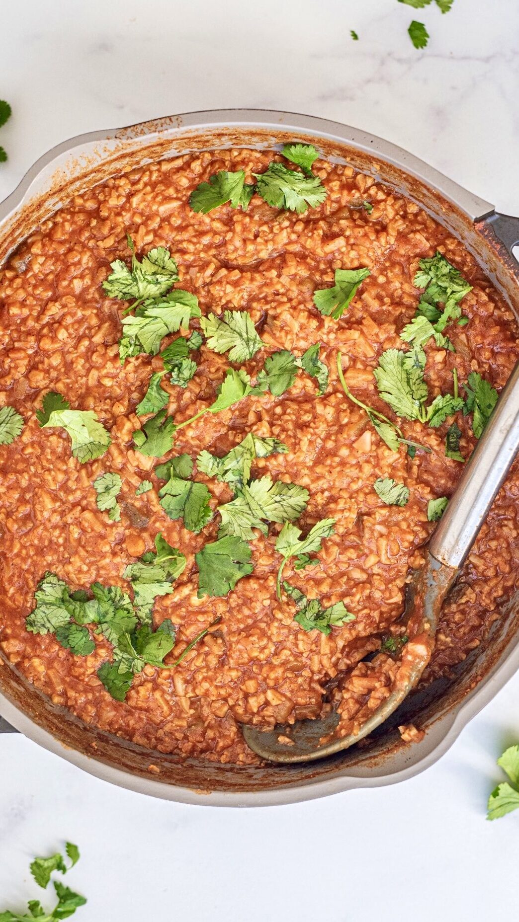 One Pan Creamy Mexican Cauliflower Rice garnished with fresh cilantro in a large pot with a silver spoon in it. Around the pot is cilantro leaves laying on the counter.