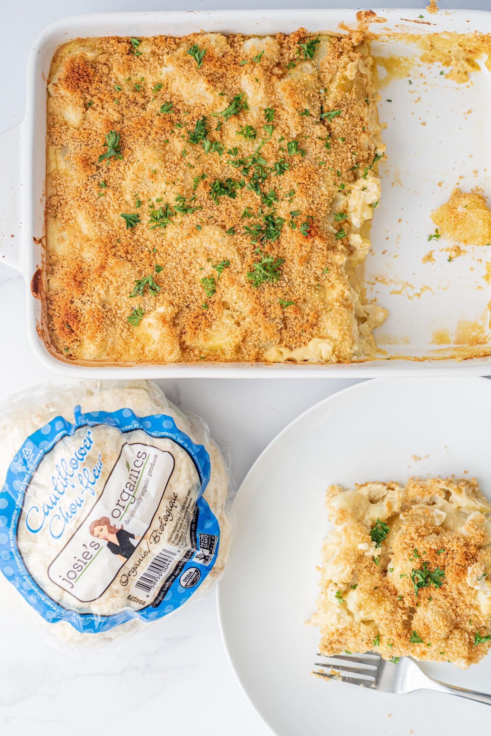 A casserole dish filled in half with a cauliflower and potato cheesy casserole with breadcrumbs and parsley on top. Beside the casserole dish is a white plate with a serving of casserole and a metal fork, and then a head of cauliflower.