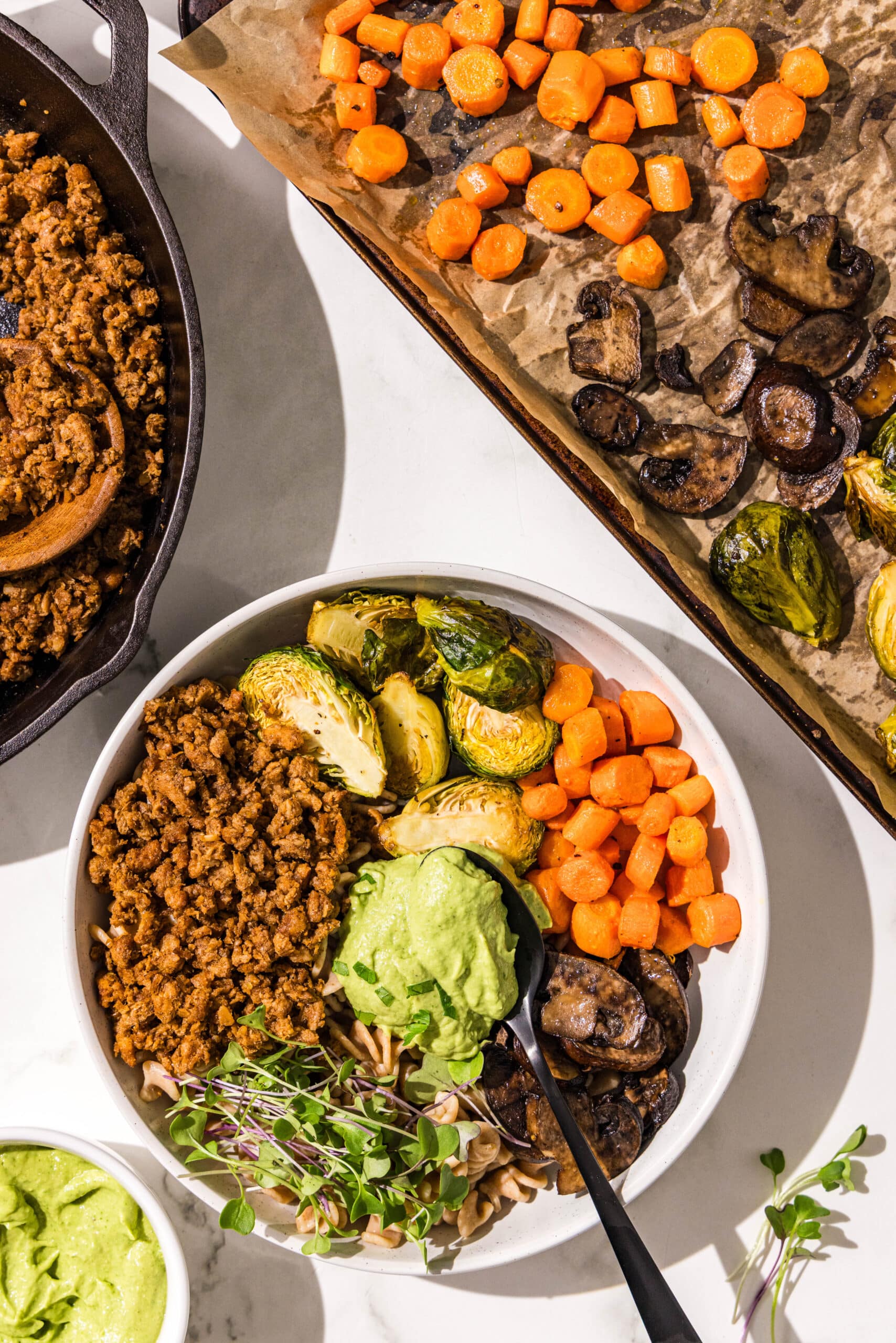 A white bowl with brussel sprouts, carrots, mushrooms, microgreens, fusilli pasta, and plant based ground beef with a scoop of green goddess sauce in the center. Beside the bowl is a cast iron pan of plant based ground beef, a sheet pan lined with parchment with carrots, mushrooms, and brussels sprouts on it, and a small bowl of avocado green goddess dressing.