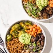 Two hormone healthy pasta bowls in white bowls. They are filled with plant based ground beef, brussels sprouts, carrots, mushrooms, pasta, microgreens, and avocado green goddess dressing in the center. Beside the bowls is two black metal forks.