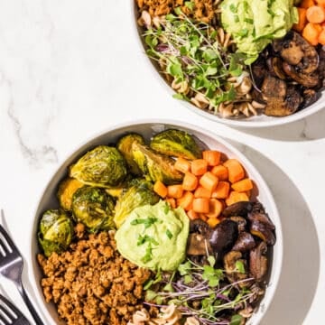 Two hormone healthy pasta bowls in white bowls. They are filled with plant based ground beef, brussels sprouts, carrots, mushrooms, pasta, microgreens, and avocado green goddess dressing in the center. Beside the bowls is two black metal forks.