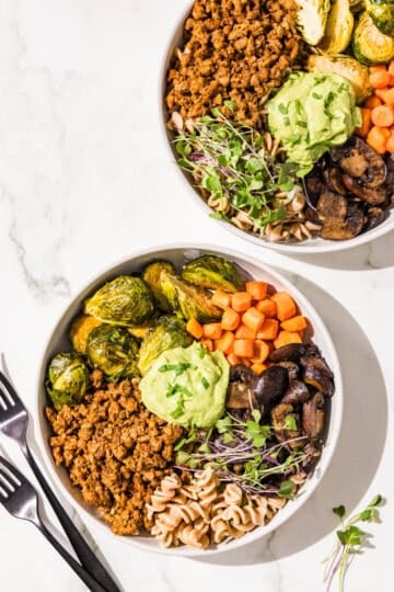 Two hormone healthy pasta bowls in white bowls. They are filled with plant based ground beef, brussels sprouts, carrots, mushrooms, pasta, microgreens, and avocado green goddess dressing in the center. Beside the bowls is two black metal forks.