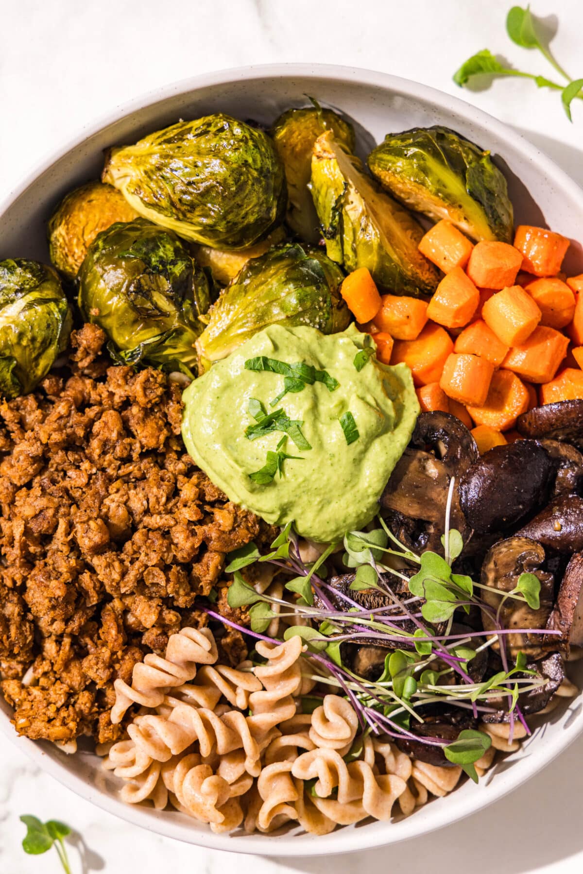 A white bowl with brussel sprouts, carrots, mushrooms, microgreens, fusilli pasta, and plant based ground beef with a scoop of green goddess sauce in the center.