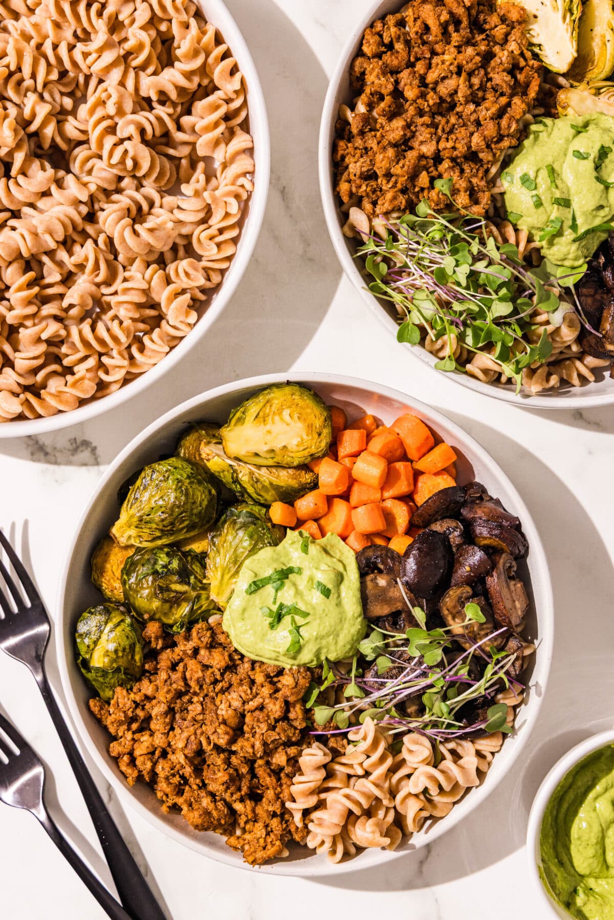 Two hormone support pasta bowls with a third bowl next to them filled with remaining plain fusili pasta noodles. Beside the bowls is a set of two black metal forks and a small bowl filled with green sauce.