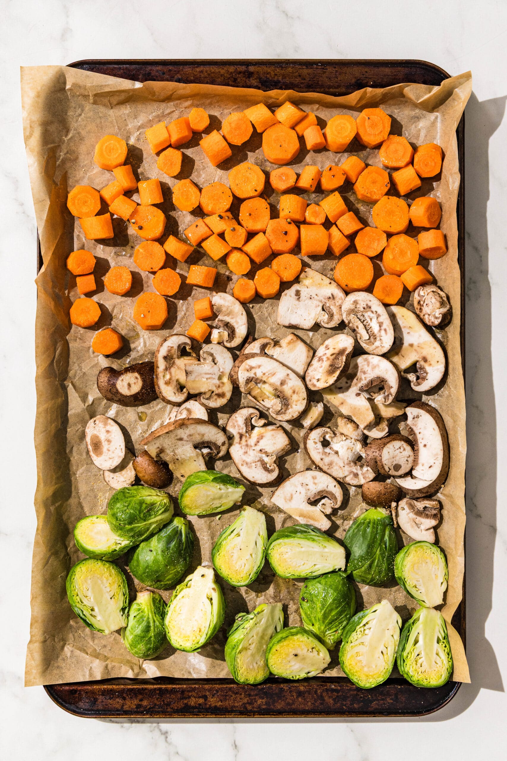 A sheet pan lined with parchment paper filled with chopped carrots, sliced mushrooms, and halved brussels sprouts.