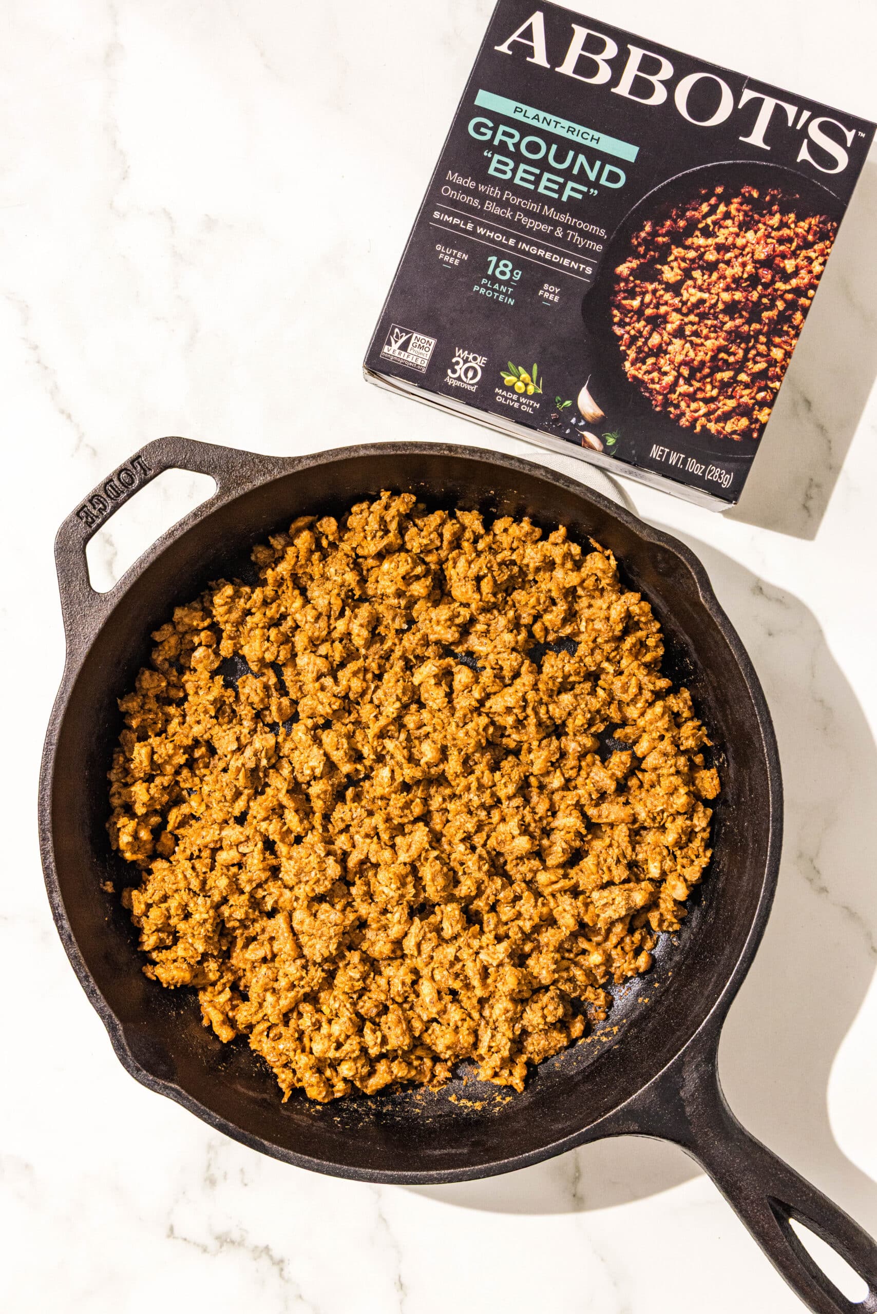 A cast iron pan filled with plant based ground beef. Beside the pan is a small black box labeled "ABBOTS" with a description of "plant based ground beef" on it.