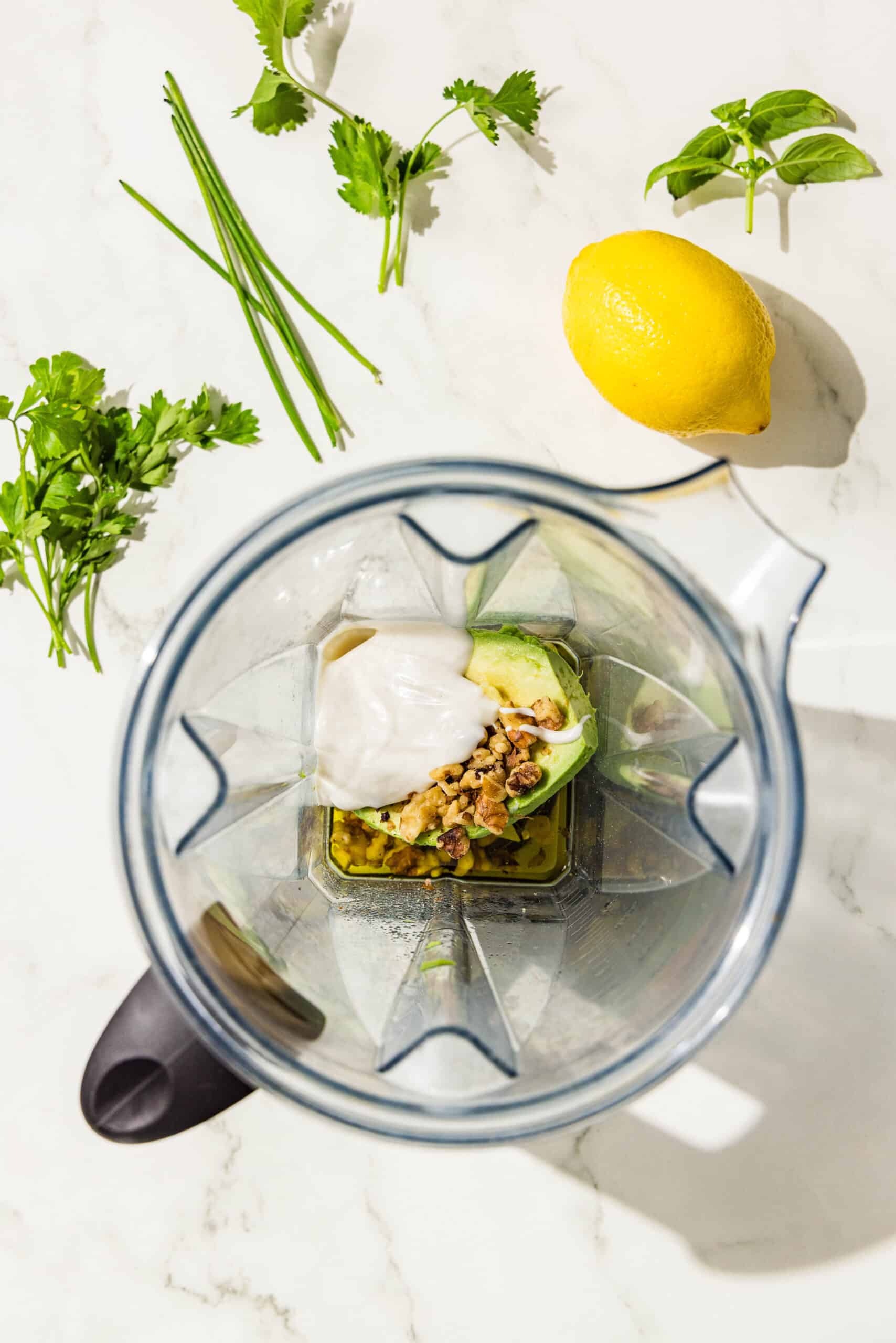 A blender filled with avocado, yogurt, walnuts, and olive oil. Beside the blender on the counter is a lemon, chives, sprig of basil, parsley, and cilantro.
