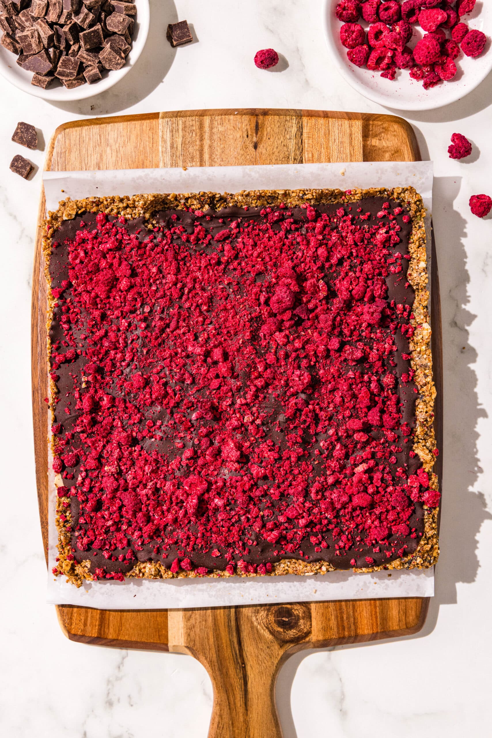 A wooden cutting board covered in parchment paper with the batch of dark chocolate raspberry cereal bars on it ready to be cut. Beside the cutting board is two small bowls of freeze dried raspberries and dark chocolate.