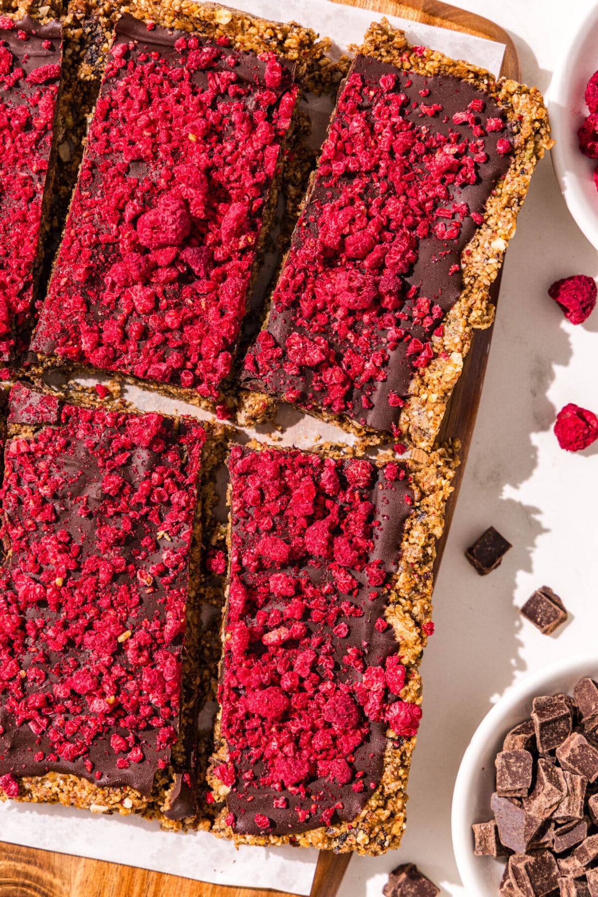 A wooden cutting board covered with parchment paper and a batch of dark chocolate raspberry cereal bars sliced. Around the cutting board is two small bowls of dried raspberries and chocolate chips.